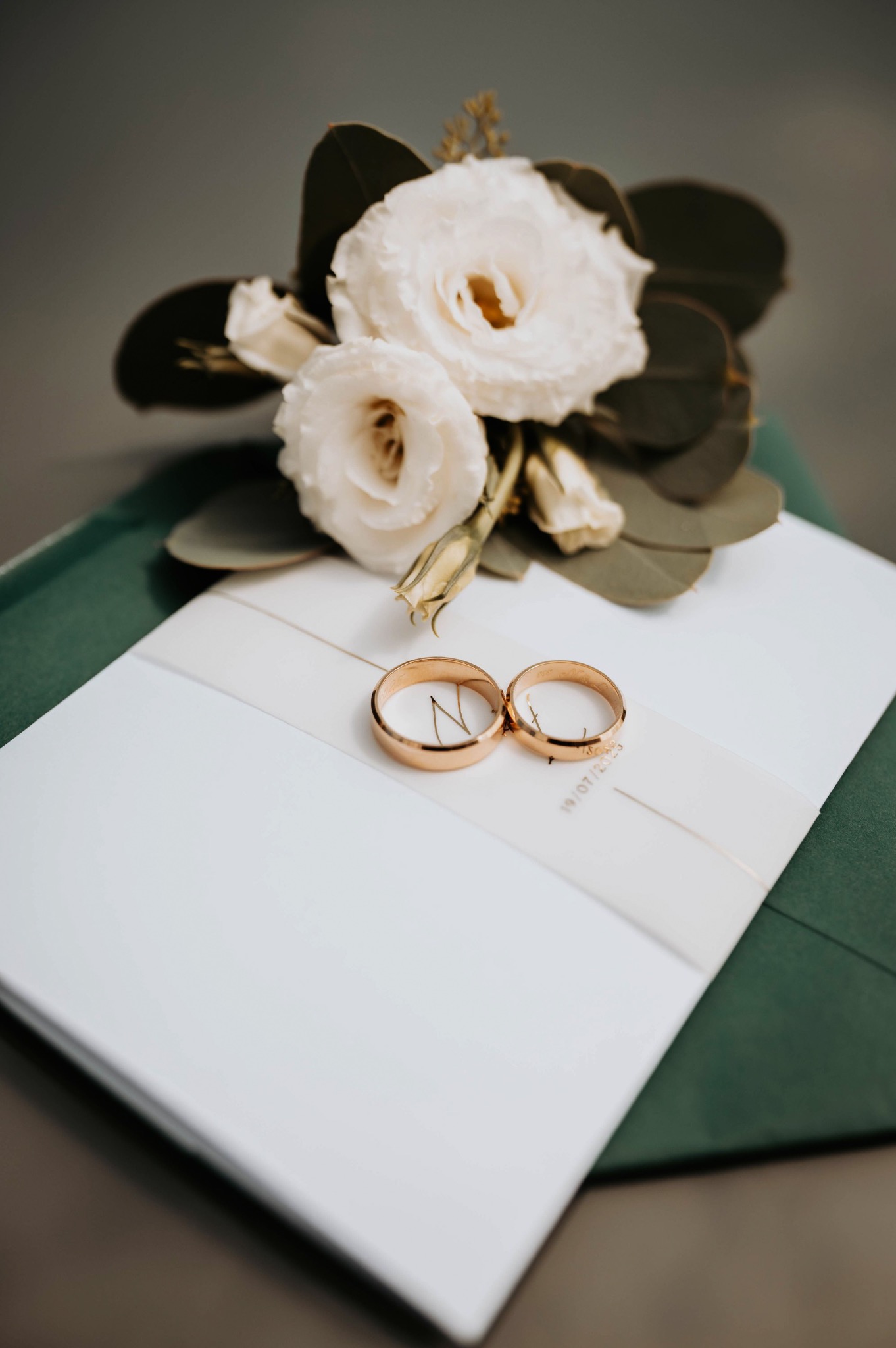 Dwa złote pierścionki ślubne leżące na białej wstążce, która owija zieloną papeterię. U góry kompozycja z białych kwiatów i zielonych liści eukaliptusa.