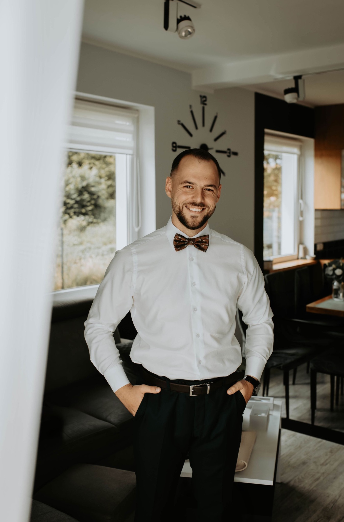 Uśmiechnięty mężczyzna w białej koszuli i muszce, z rękami w kieszeniach, pozuje w jasnym wnętrzu z zegarem na ścianie i widokiem na zieleń za oknem.