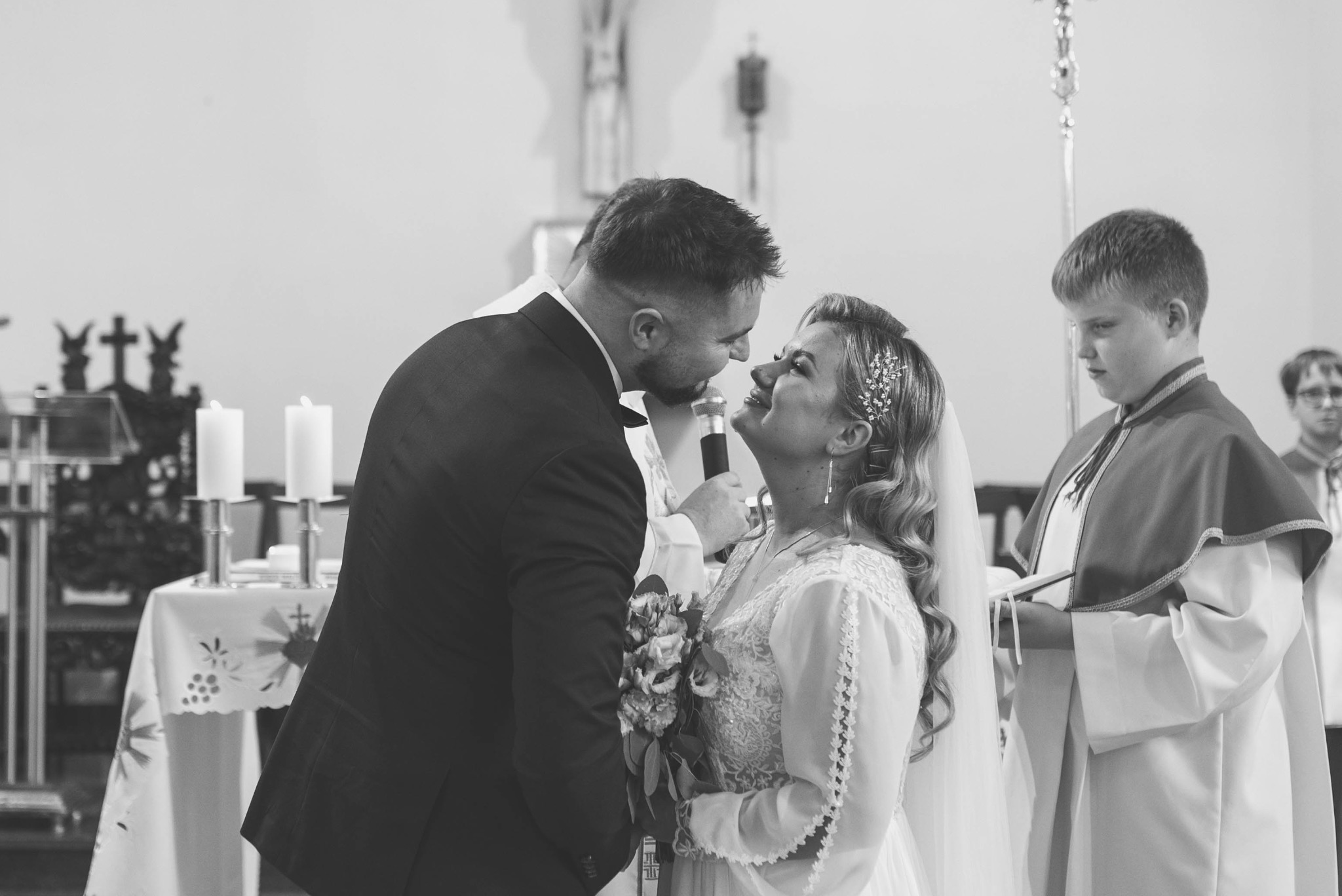 Czarno-białe ujęcie pary młodej podczas ceremonii ślubnej w kościele, z ministrantem w tle. Panna młoda trzyma bukiet, a pan młody mówi do mikrofonu.