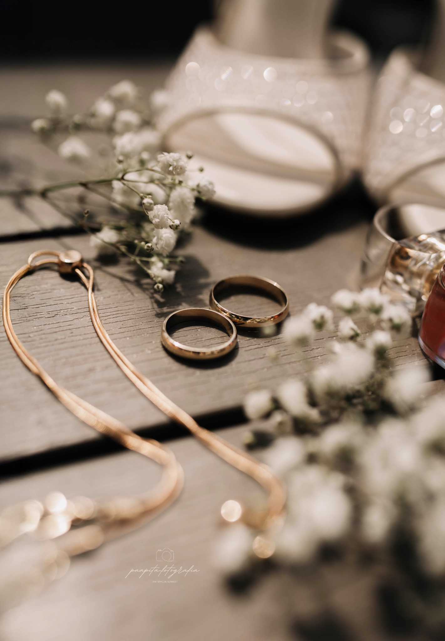 Dwa złote obrączki ślubne na drewnianym tle, otoczone drobnymi białymi kwiatami i elementami biżuterii, z delikatnym oświetleniem i butami ślubnymi w tle.