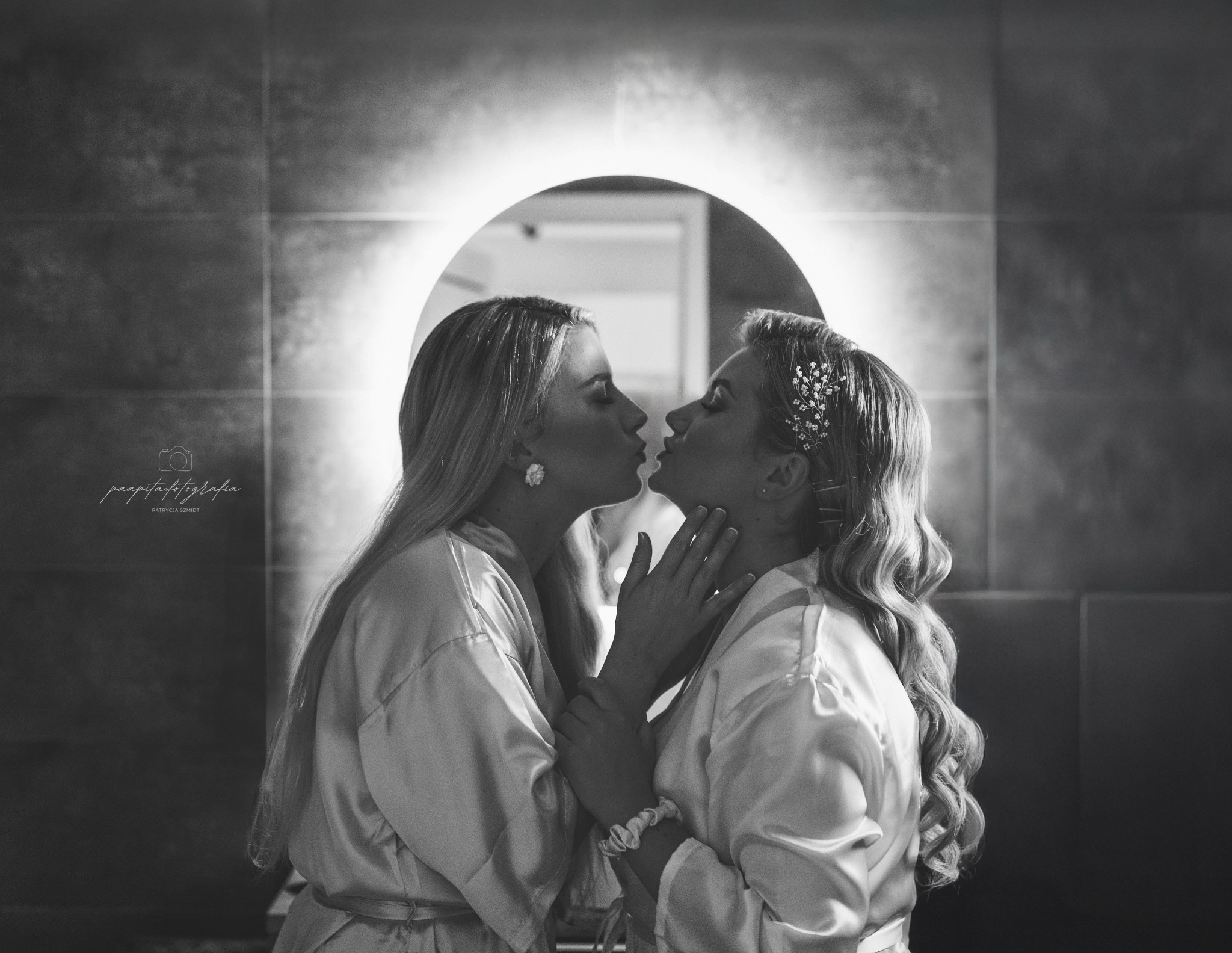 Czarno-białe zbliżenie dwóch kobiet w szlafrokach, delikatnie się całujących na tle podświetlonego lustra. Jedna z ozdobą we włosach, druga z kwiatowym kolczykiem.