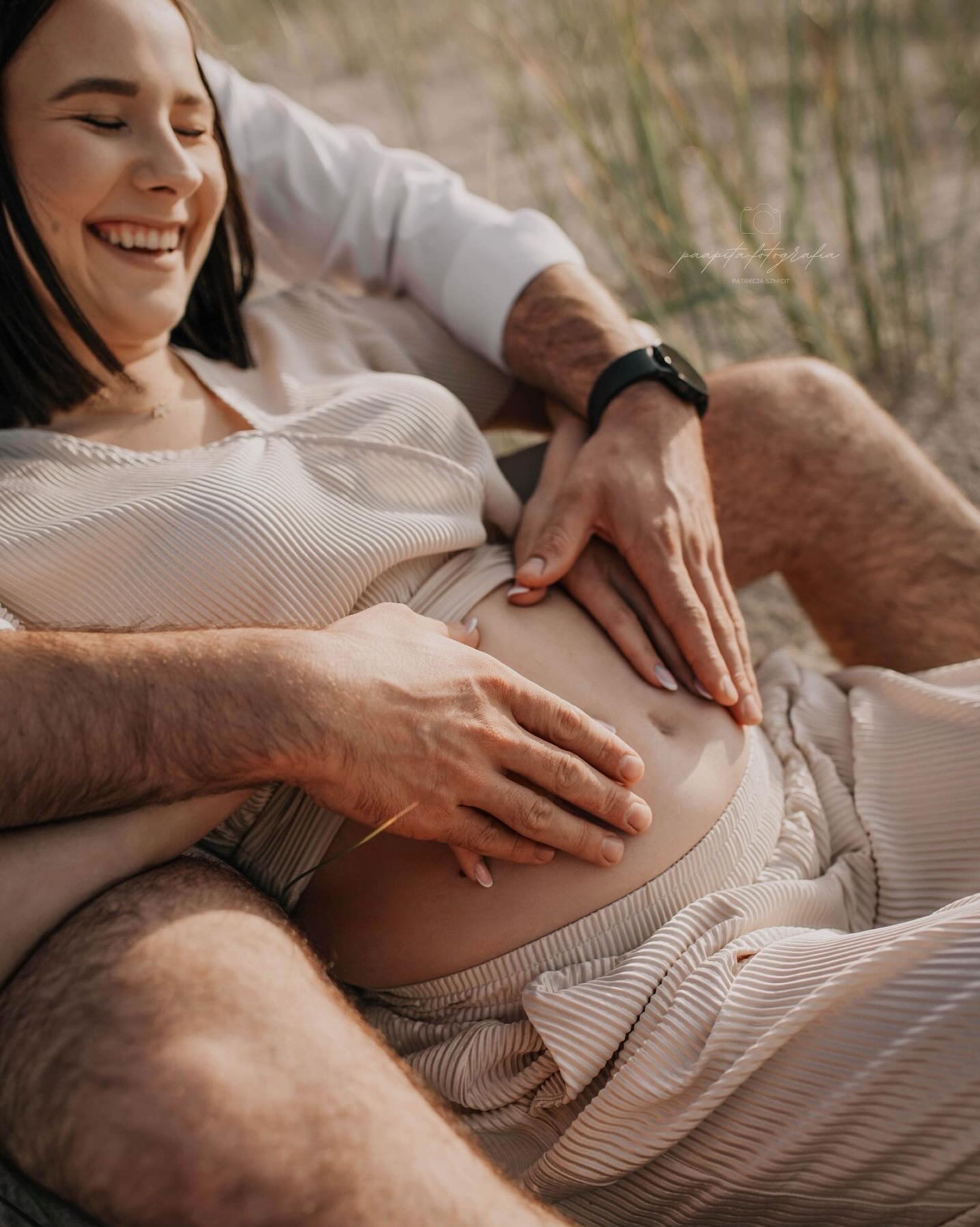 Uśmiechnięta przyszła mama z partnerem na plaży, dłonie na brzuchu, ciepłe światło, naturalna sceneria, radosne oczekiwanie, delikatna kolorystyka.