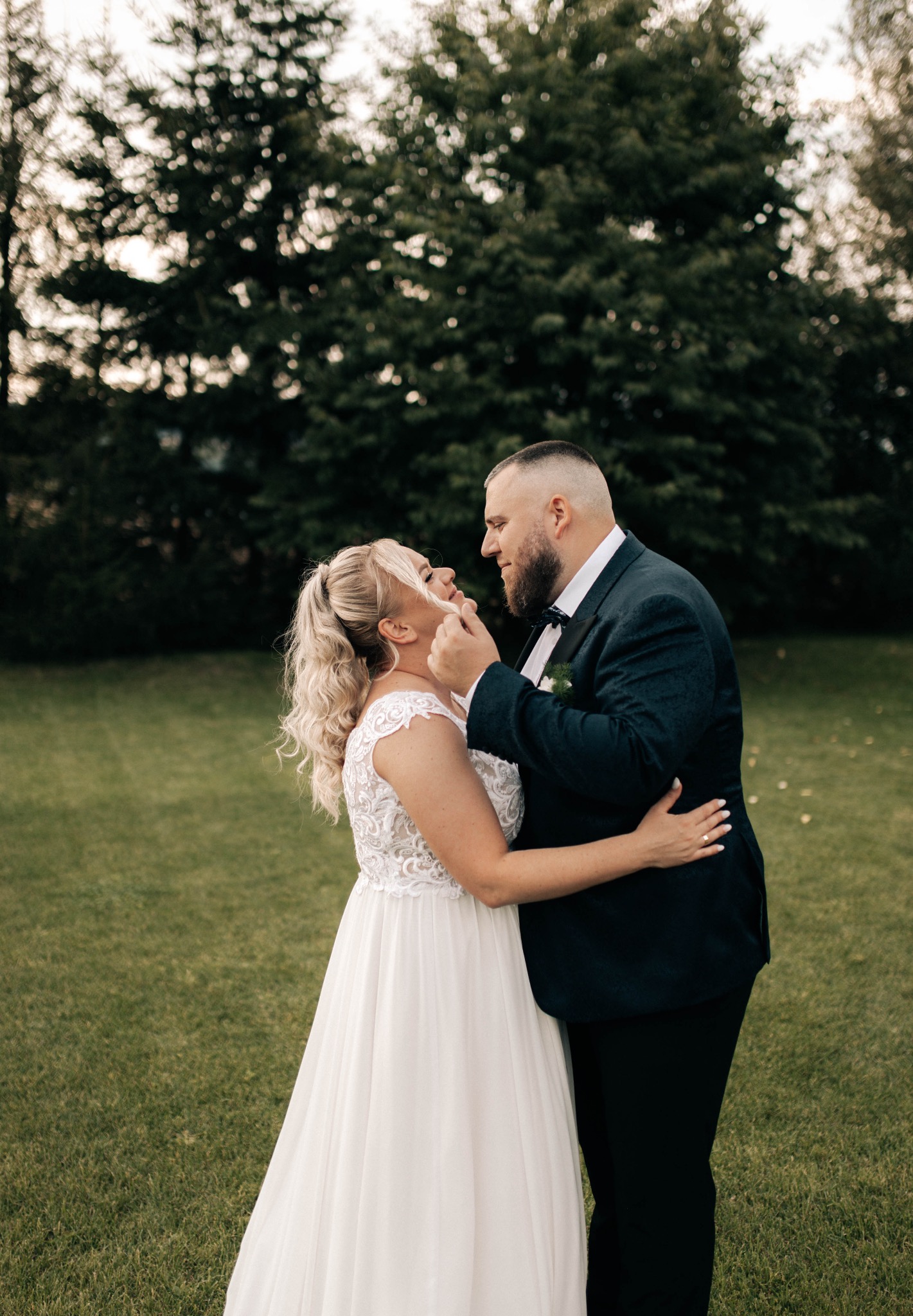 Para młoda w objęciach na tle zielonego trawnika i drzew. Panna młoda w białej sukni z koronkową górą, pan młody w ciemnym garniturze z muszką.