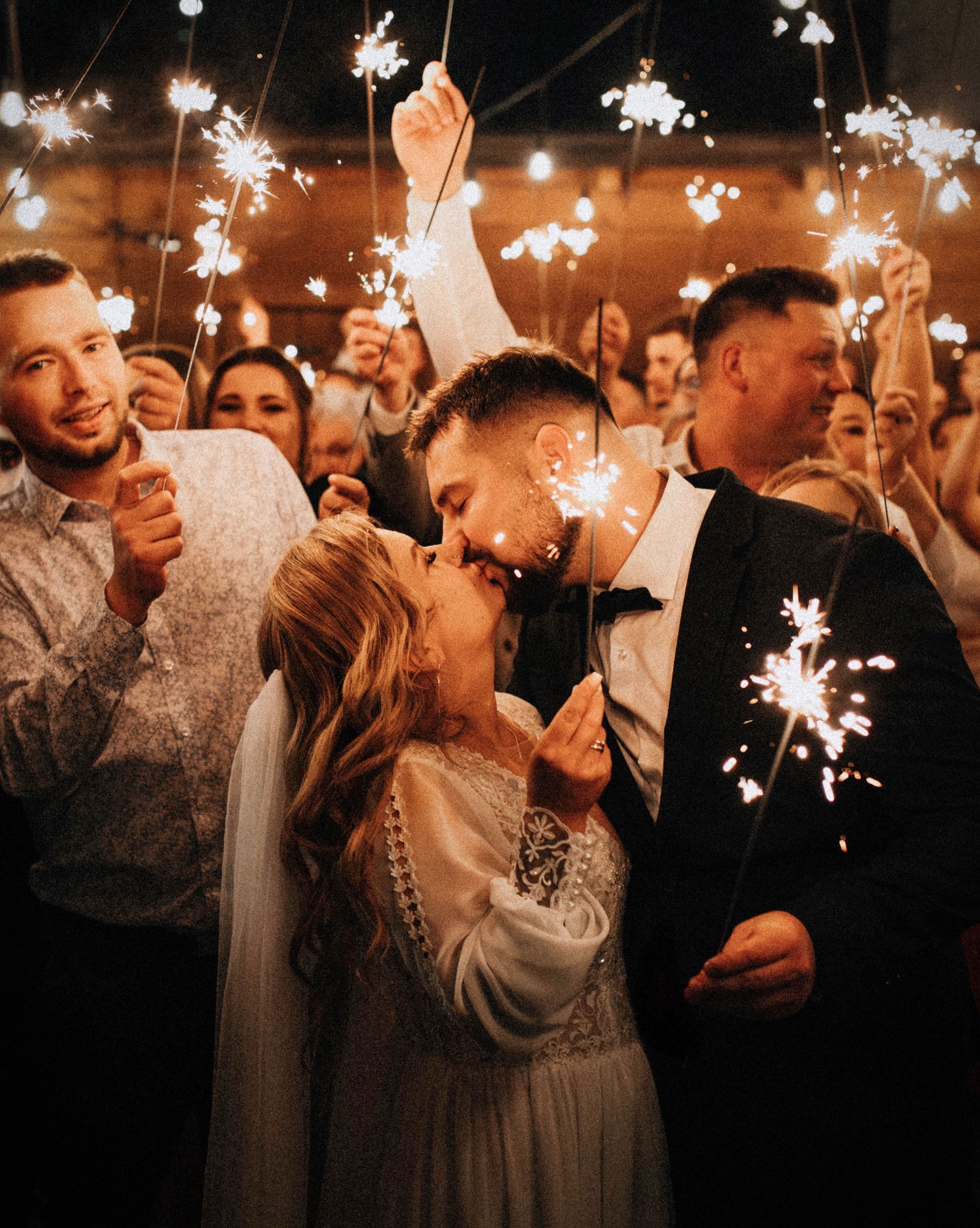 Pocałunek pary młodej otoczonej gośćmi z zimnymi ogniami, ciepłe światło, uroczysta atmosfera, suknia ślubna z koronkowymi detalami, czarny smoking.