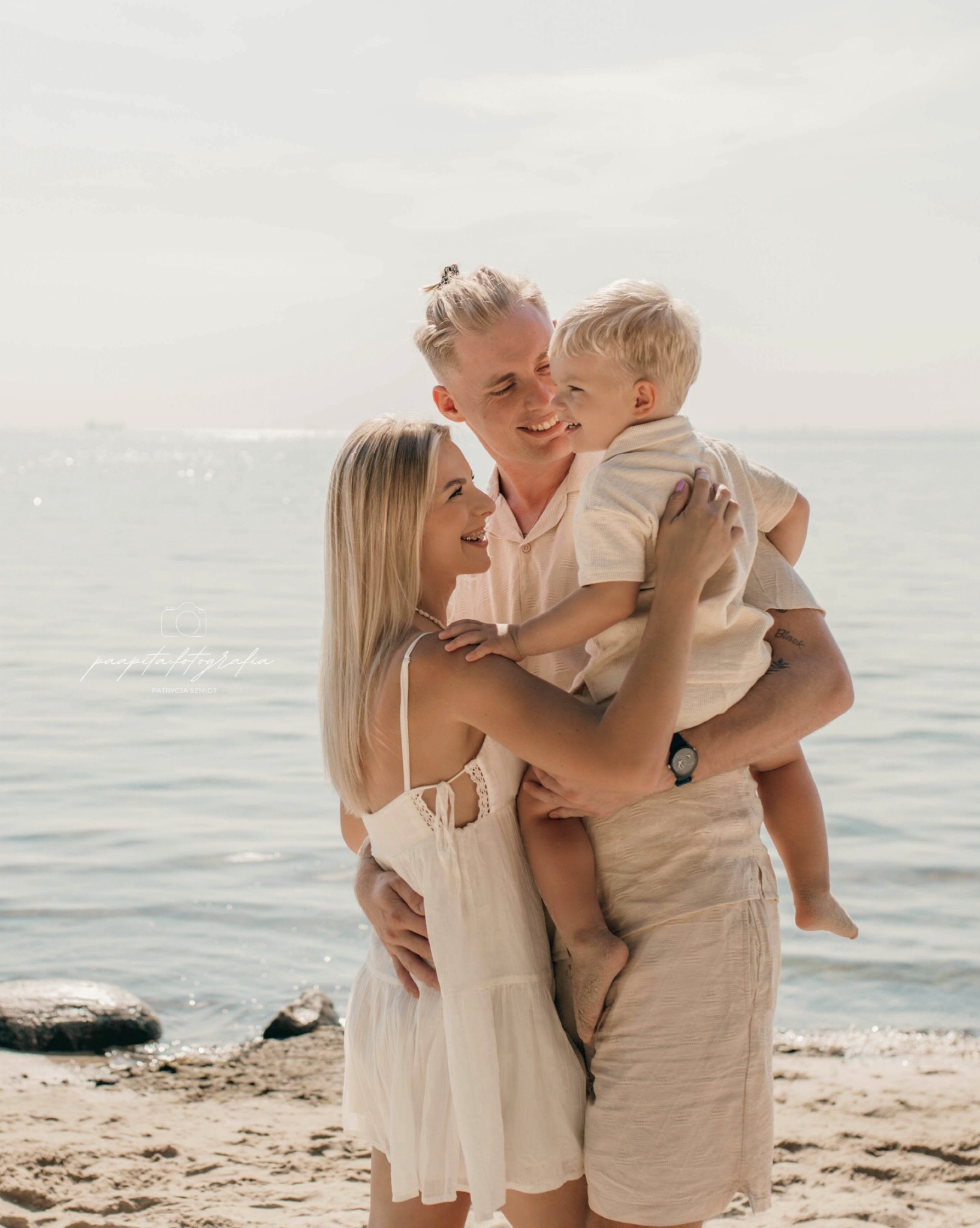 Uśmiechnięta rodzina na plaży w jasnych ubraniach: mama, tata trzymający synka. Delikatne światło, morski krajobraz w tle. Sesja rodzinna w naturalnym stylu.