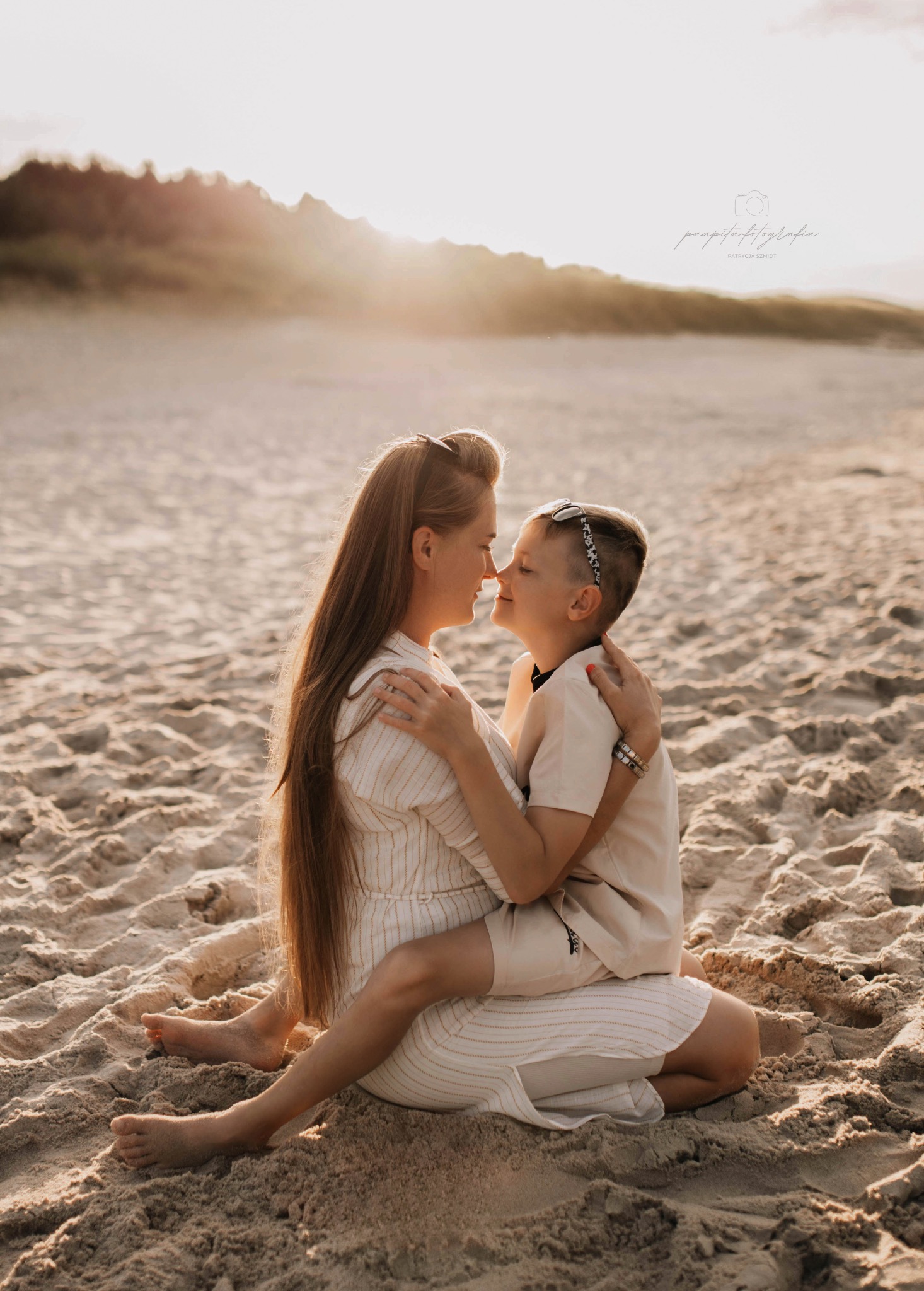 Matka z synem siedzą na piasku, dotykając się nosami w ciepłym, zachodzącym słońcu. Ubrani w jasne stroje, emanują bliskością i radością. Romantyczna fotografia rodzinna.