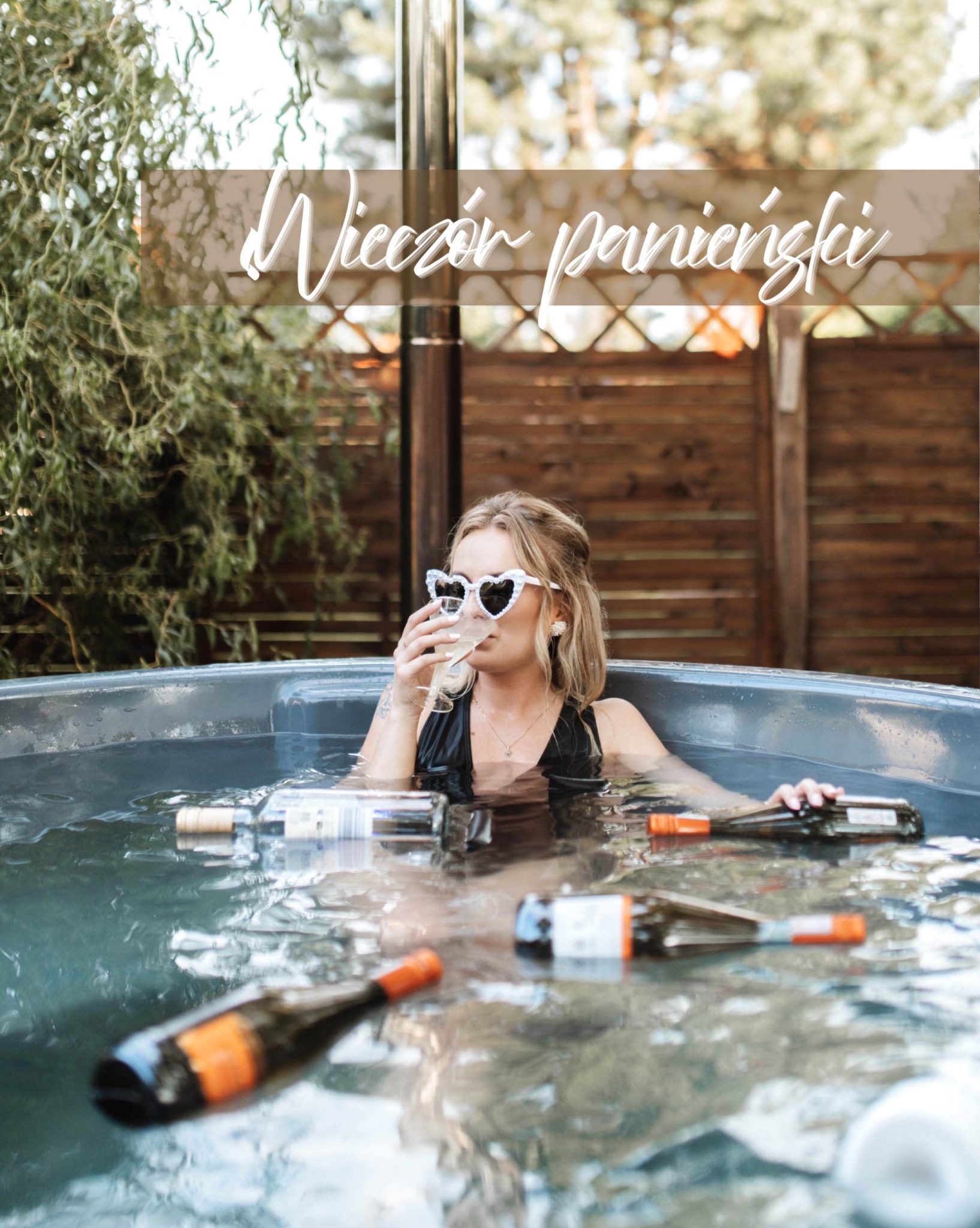 Kobieta w okularach w kształcie serc relaksuje się w jacuzzi z butelkami wina. Ujęcie z wieczoru panieńskiego na tle drewnianego ogrodzenia i zieleni.