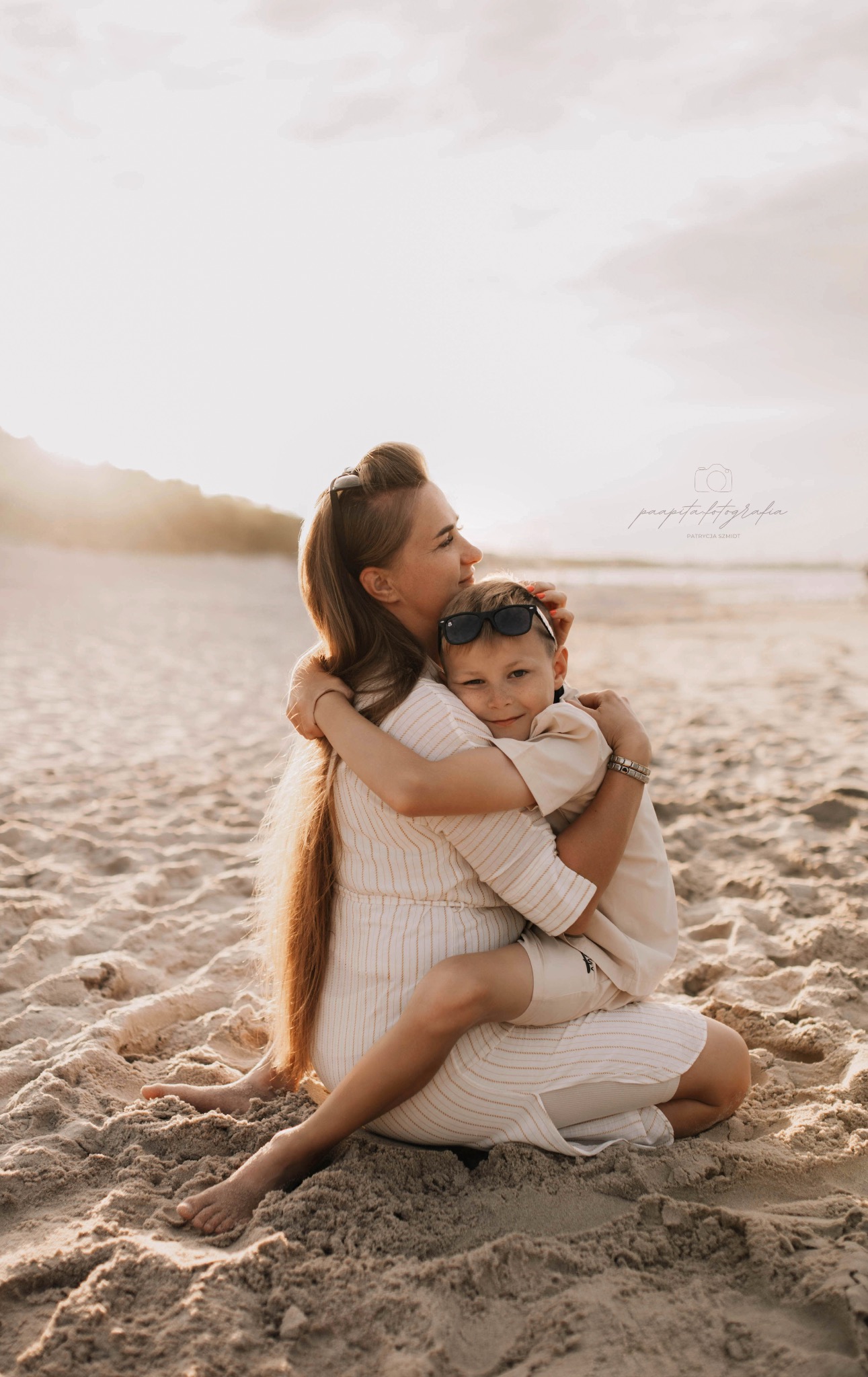 Mama tuli syna w okularach na plaży o zachodzie słońca. Ciepłe barwy, miękkie światło, naturalna sceneria. Uśmiechnięty chłopiec, bliskość i miłość w kadrze.