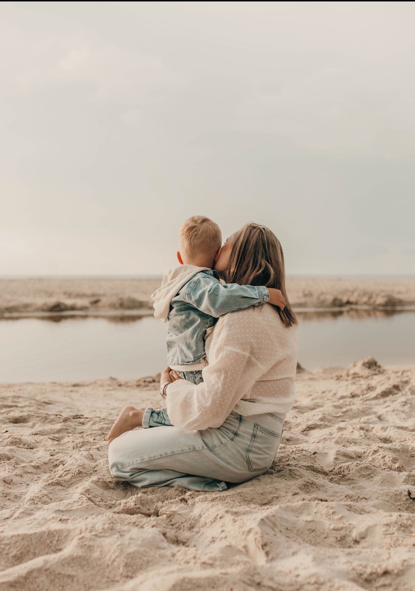 Matka siedzi na piasku, obejmując syna, który ją całuje w policzek. W tle woda i niebo w pastelowych barwach. Ujęcie z tyłu, delikatne światło.