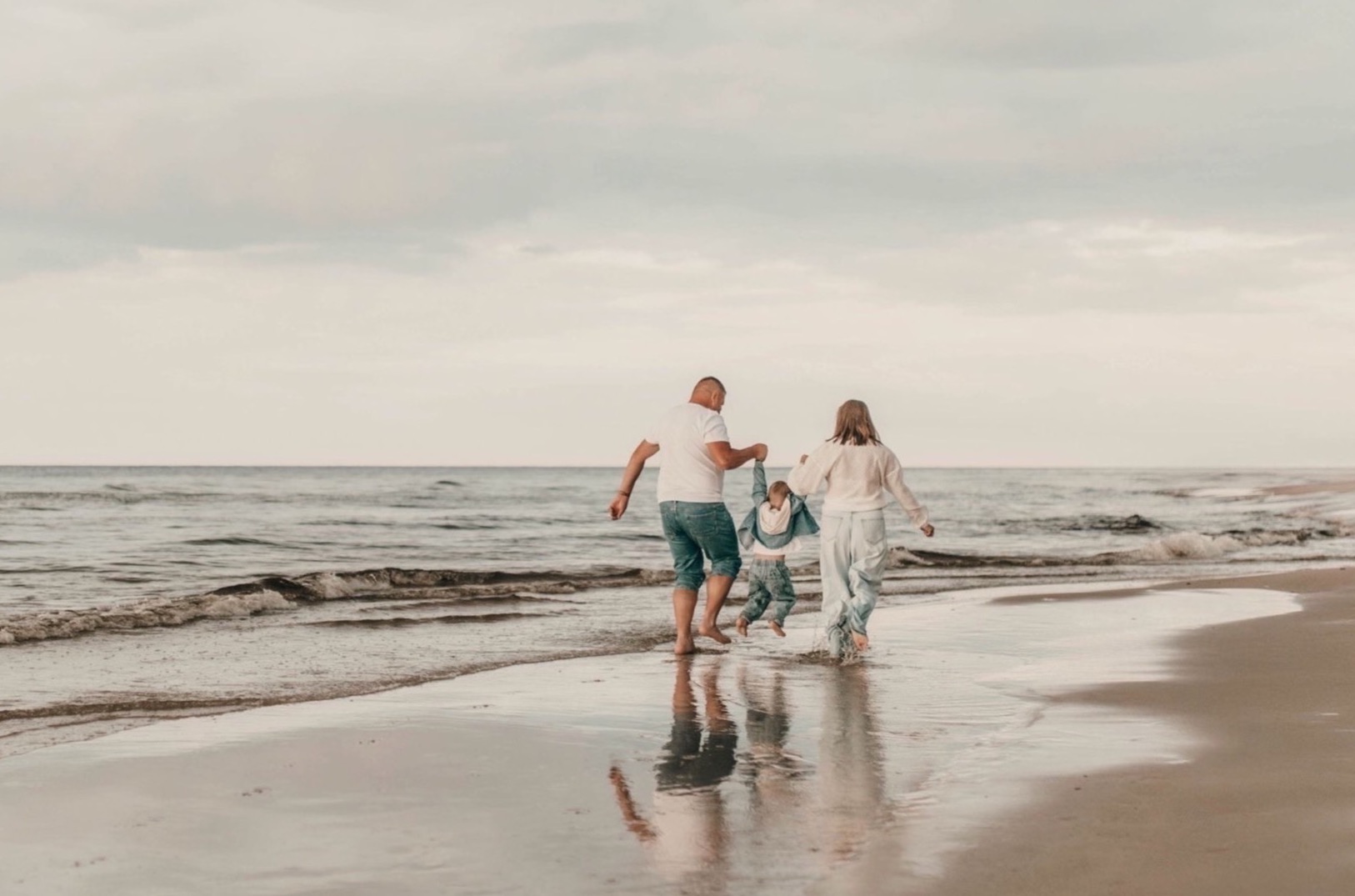 Rodzinna sesja na plaży: rodzice trzymają za ręce dziecko, biegnąc brzegiem morza. Delikatne kolory, odbicie w wodzie, spokojna atmosfera.