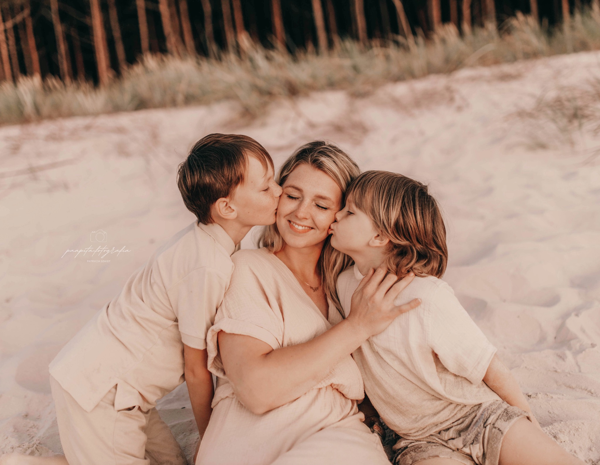 Mama z dwoma synami na plaży, obaj całują ją w policzki. Ubrani w beżowe ubrania, delikatne światło, naturalna sceneria z drzewami w tle. Ciepła, rodzinna atmosfera.