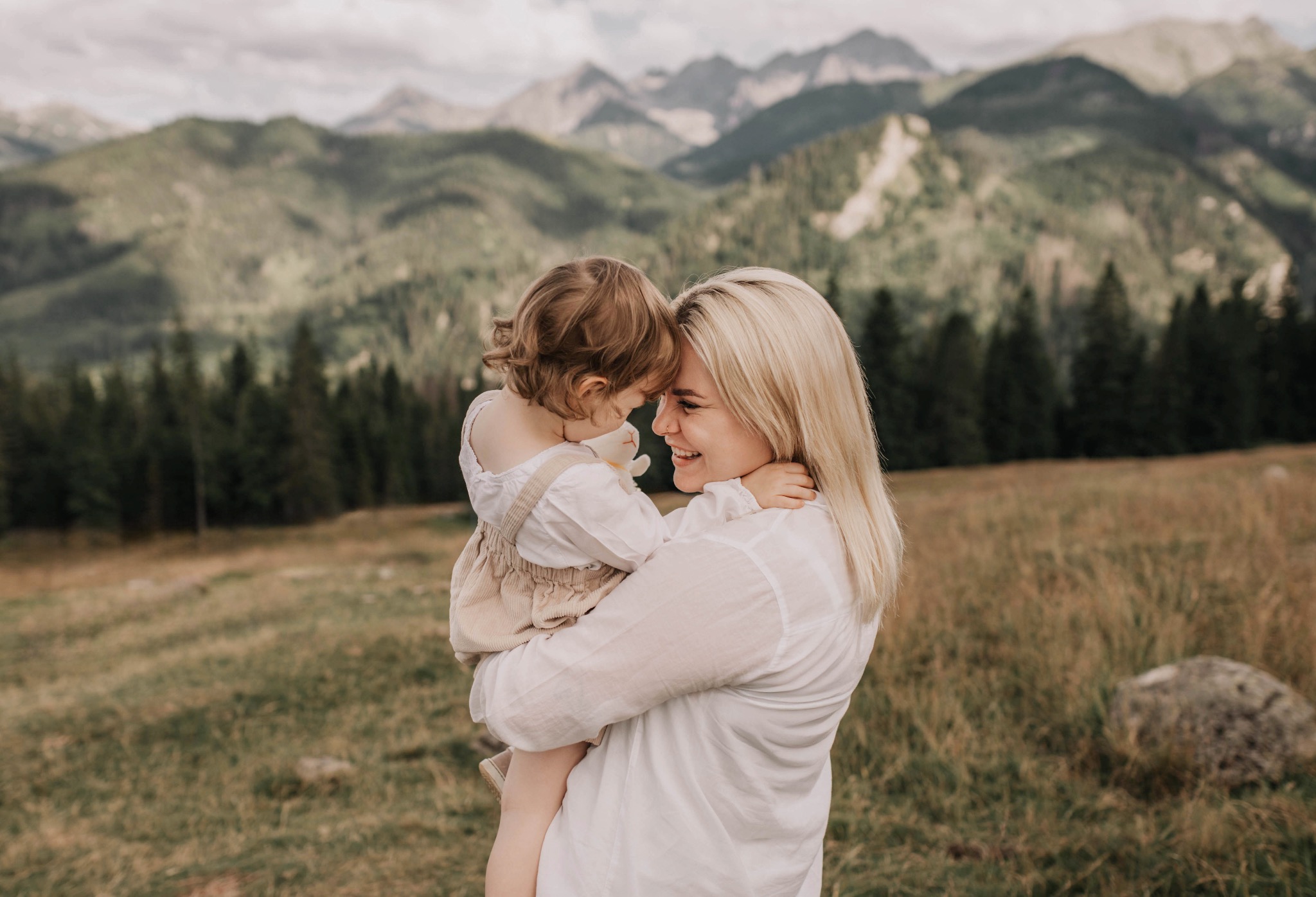 Uśmiechnięta blond mama tuli córeczkę na tle gór. Romantyczna sesja rodzinna w plenerze, naturalne światło, ciepłe kolory, bliskość i miłość w obiektywie.