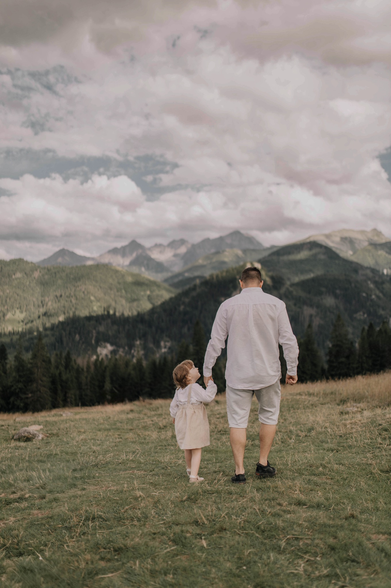 Ojciec z córką spacerują po łące na tle gór. Mężczyzna w białej koszuli i krótkich spodenkach trzyma za rękę małą dziewczynkę w sukience.