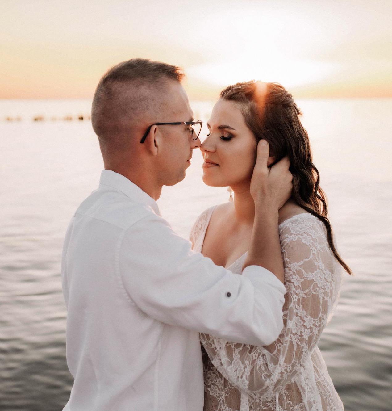 Para w białych strojach, zbliżona do pocałunku na tle zachodzącego słońca nad wodą. Delikatne światło podkreśla romantyczny nastrój.