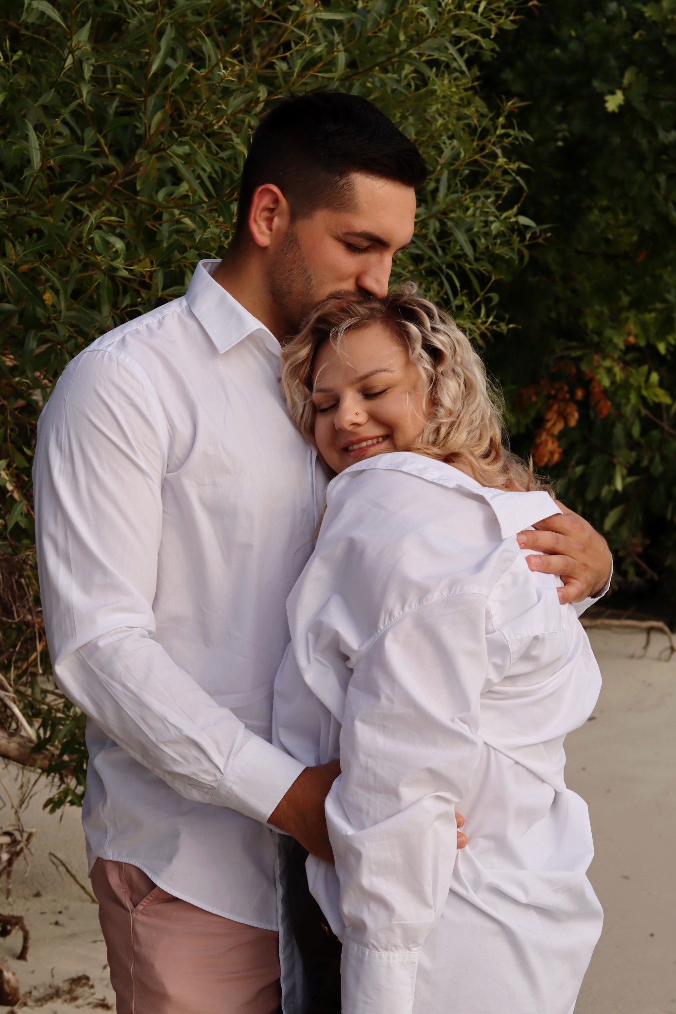 Uśmiechnięta kobieta z blond lokami w objęciach mężczyzny, który całuje ją w czoło, oboje ubrani w białe koszule na tle zieleni.