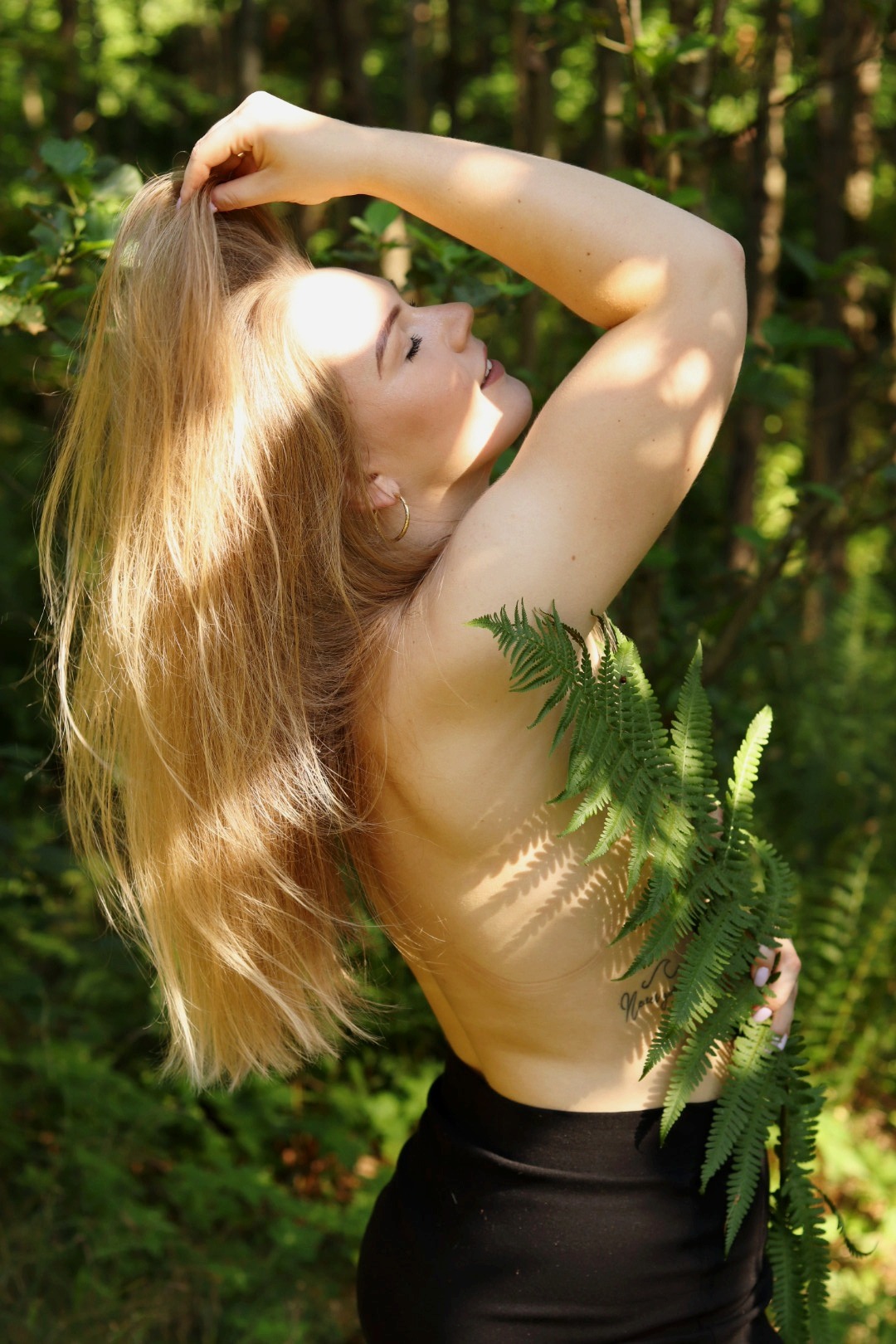 Sesja kobieca w Gniewinie: Blondwłosa modelka pozuje w naturalnym świetle, z paprocią, zmysłowo odchylona do tyłu, w otoczeniu zieleni. Subtelny tatuaż na plecach.