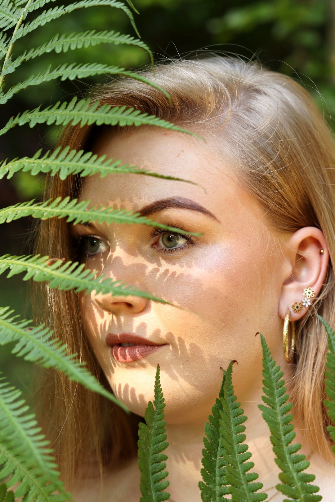 Portret młodej kobiety z blond włosami, zza paproci, z delikatnym makijażem i złotymi kolczykami w kształcie kwiatów i koła.