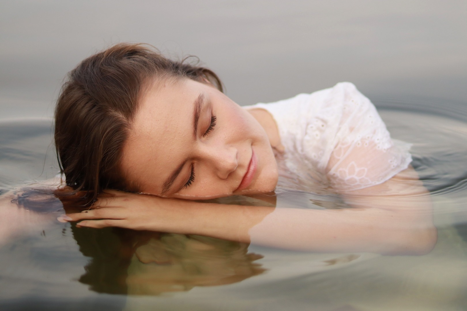 Kobieta w białej koronkowej sukience zanurzona w wodzie, leżąca na rękach z zamkniętymi oczami i delikatnym uśmiechem, odbicie w tafli wody.