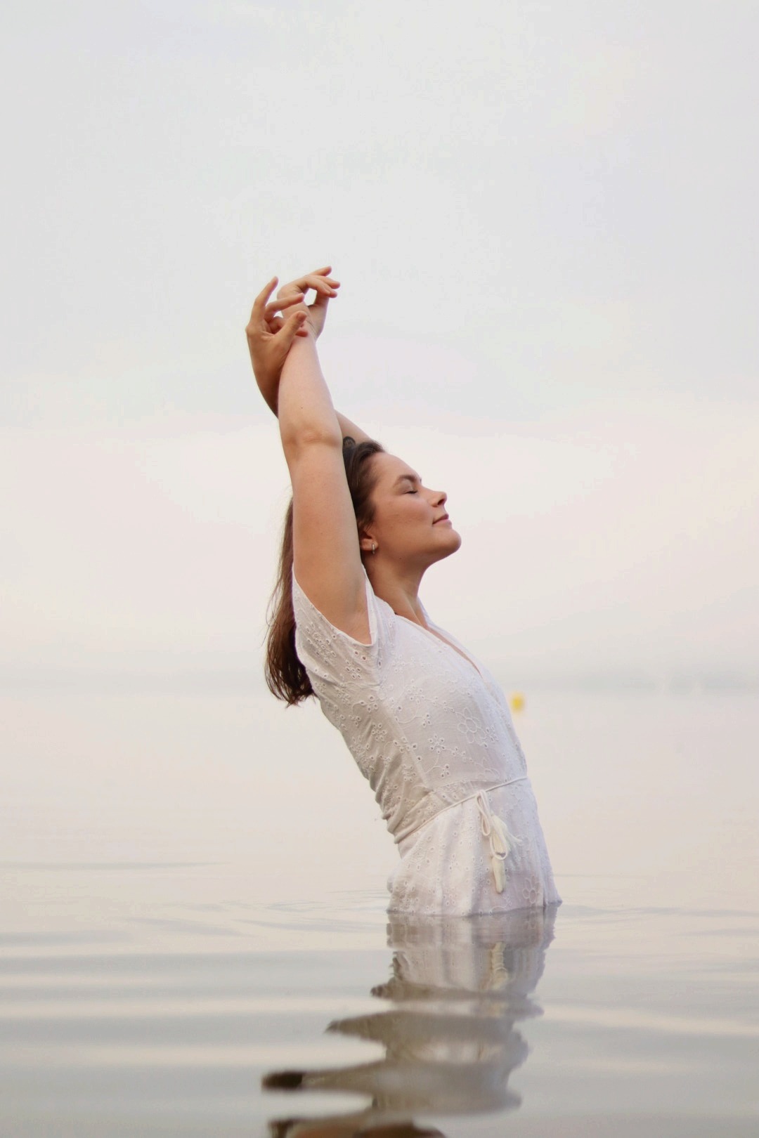 Kobieta w białej sukience zanurzona w wodzie po pas, z rękami uniesionymi do góry, odchylona do tyłu, z zamkniętymi oczami.