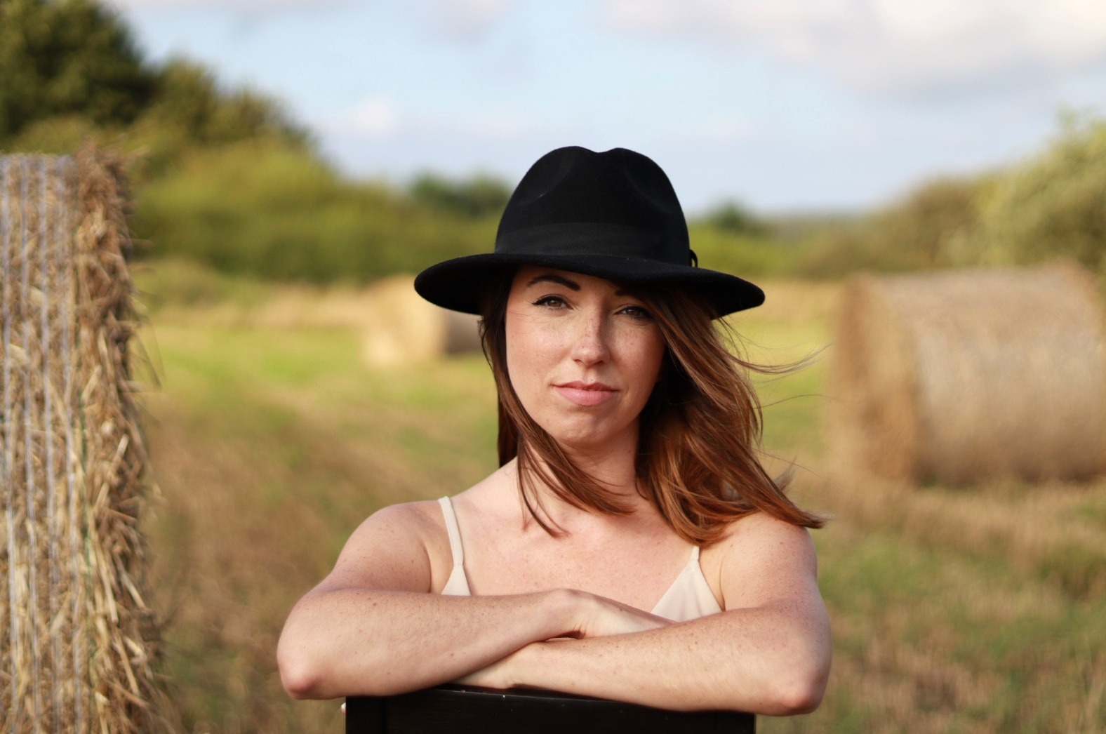 Portret kobiety w czarnym kapeluszu na tle pola ze słomianymi balotami, włosy rozwiane na wietrze.