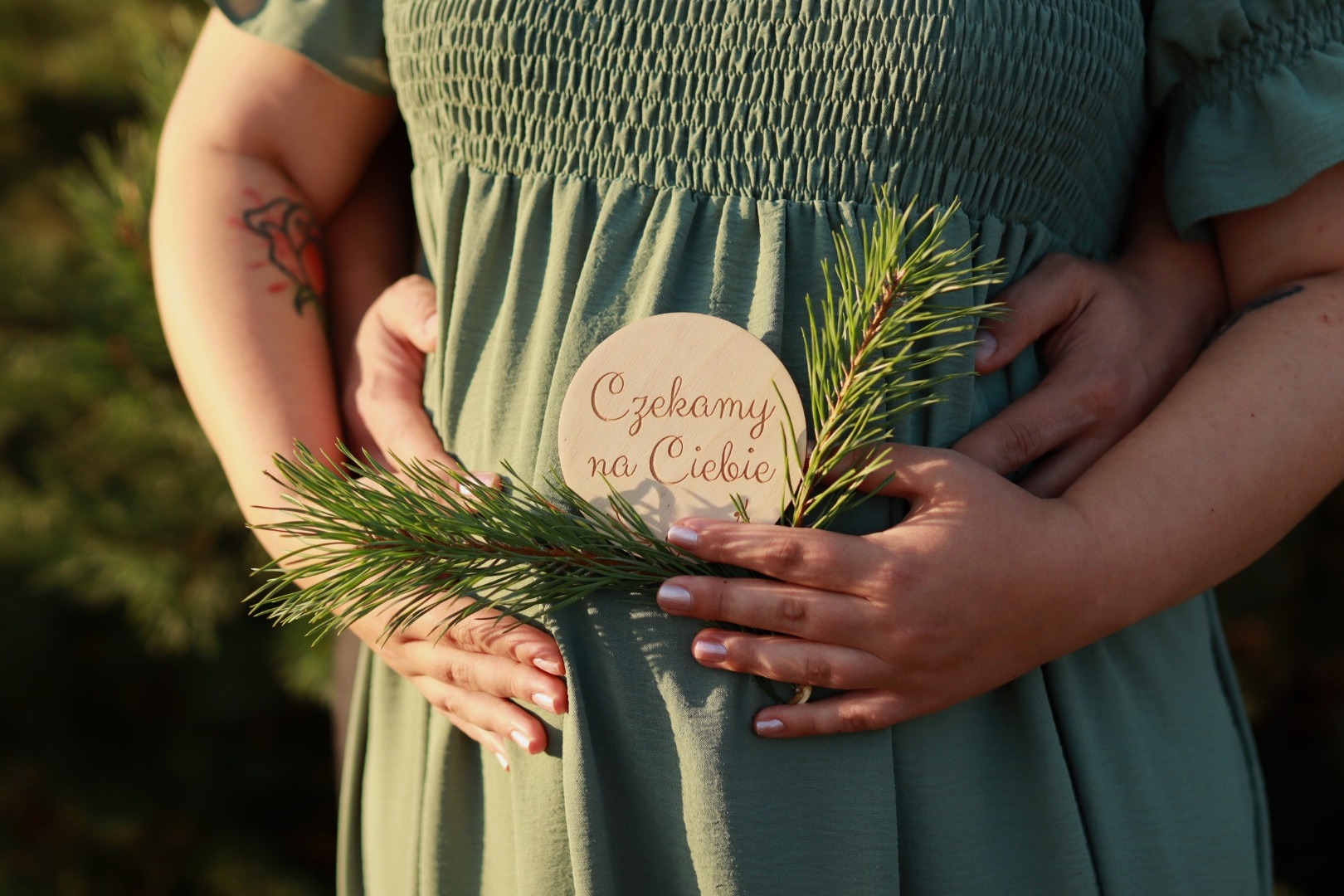Brzuch ciężarnej kobiety w zielonej sukience, trzymany przez nią i partnera, ozdobiony gałązką sosny i drewnianym krążkiem z napisem 'Czekamy na Ciebie'.
