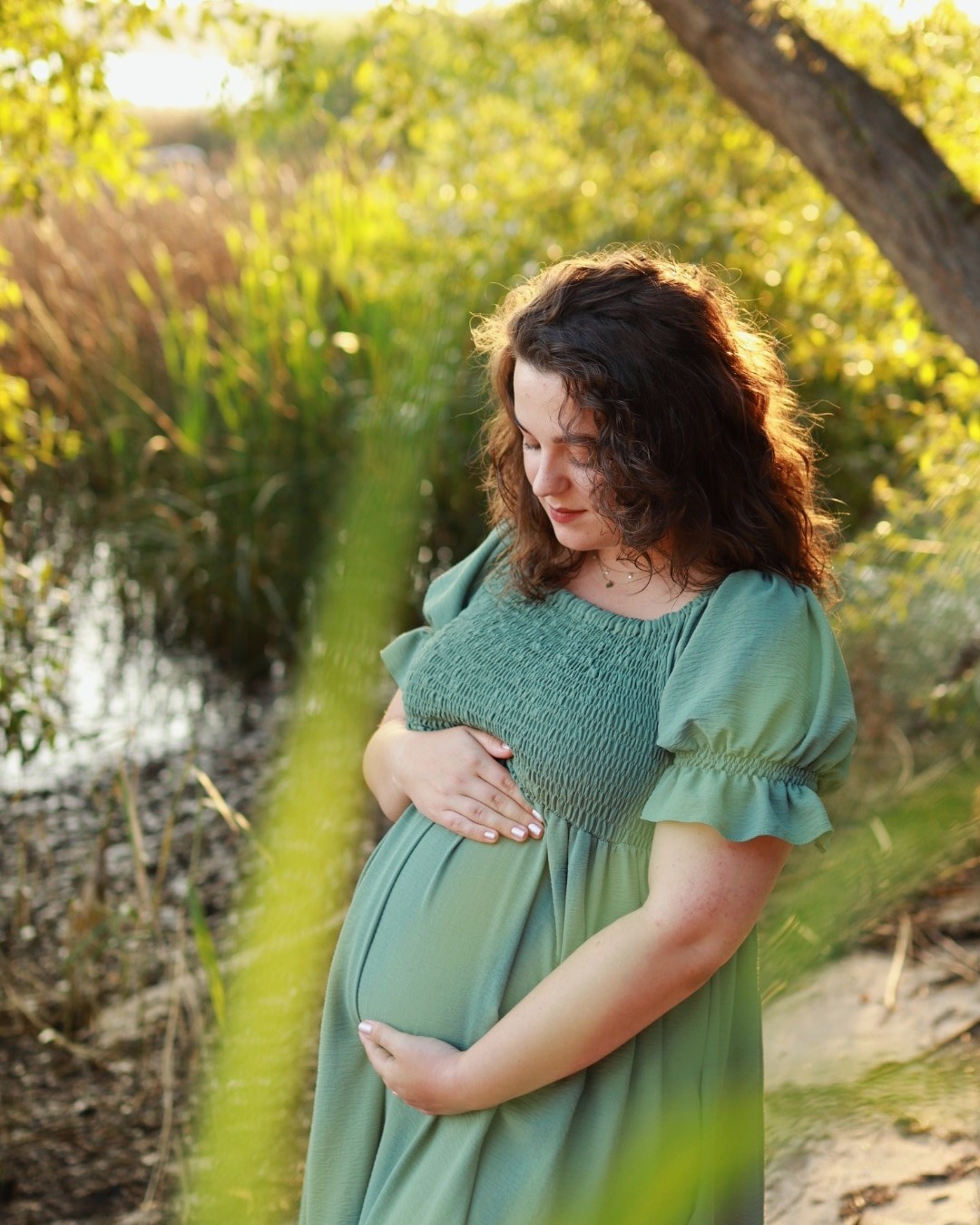 Kobieta w zielonej sukience ciążowej, z rękami na brzuchu, w zalesionym otoczeniu z wodą w tle, oświetlona ciepłym światłem popołudniowego słońca.