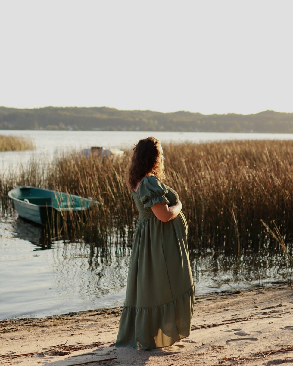 Kobieta w długiej, oliwkowej sukience ciążowej stoi na piaszczystym brzegu jeziora, z łódką i trzcinami w tle, oświetlona ciepłym światłem zachodzącego słońca.