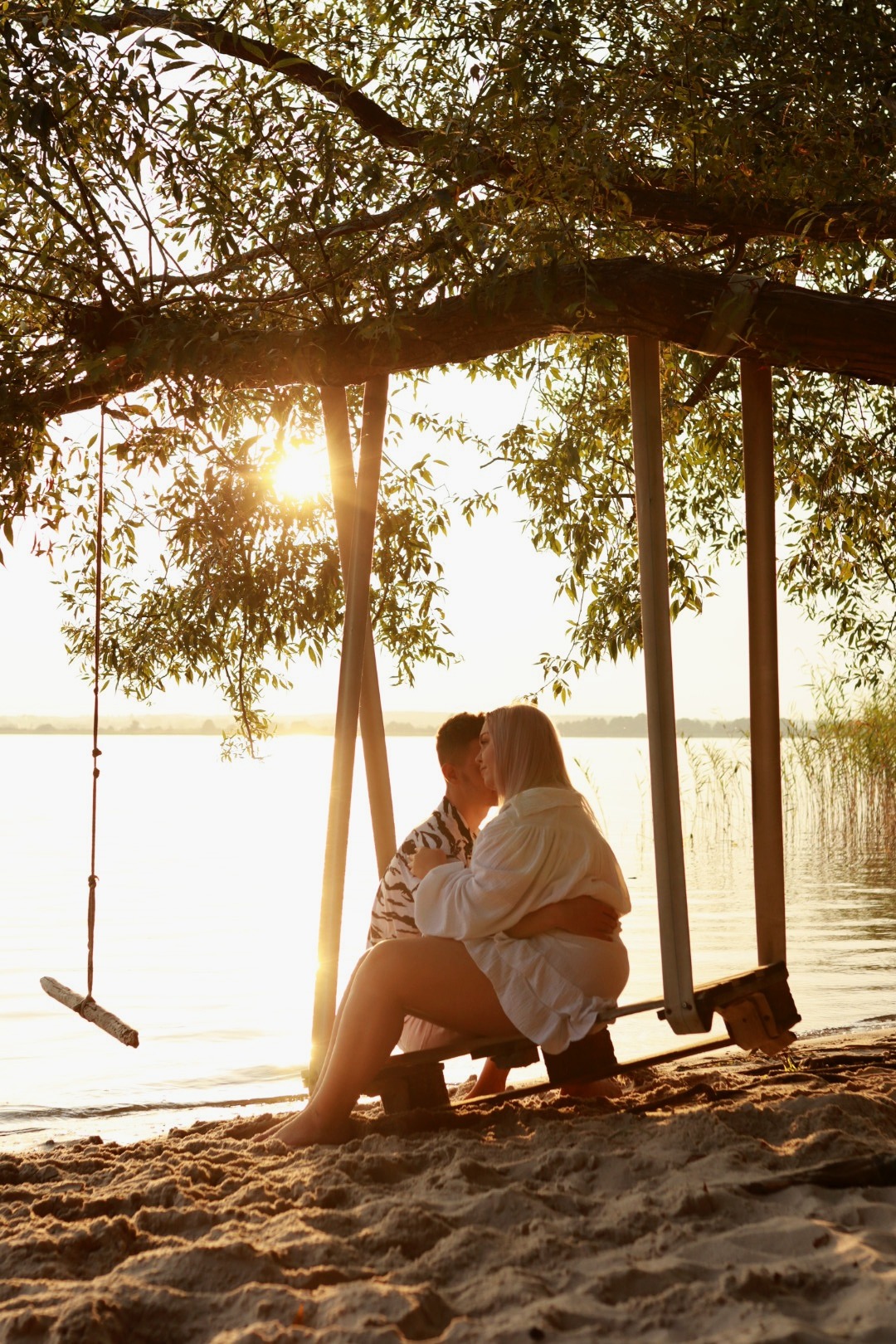 Para całująca się na drewnianej huśtawce pod drzewem na piaszczystej plaży o zachodzie słońca, widok na jezioro w tle.