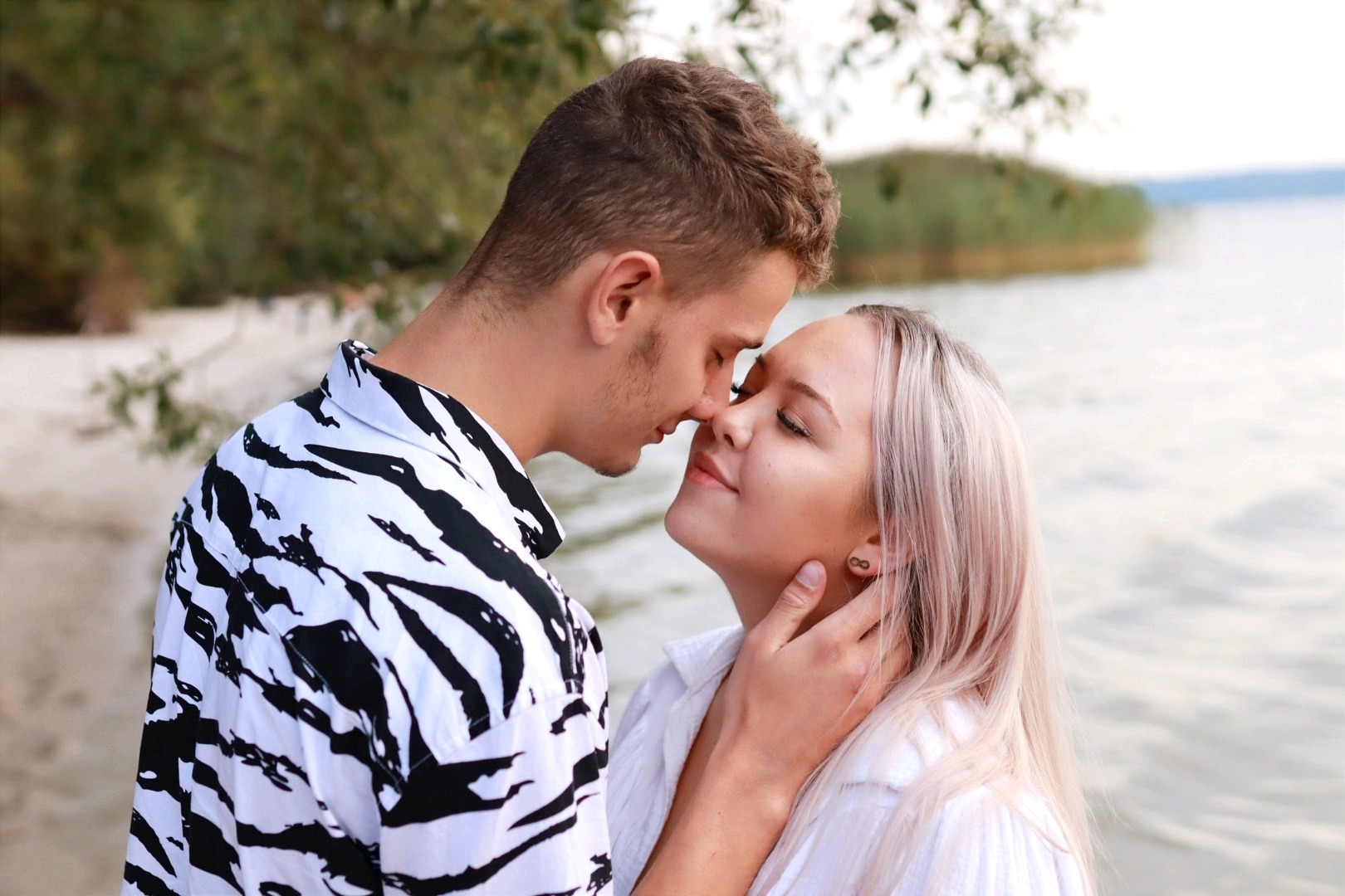 Para delikatnie dotykająca się nosami na tle jeziora, mężczyzna w koszuli w zebrę, kobieta z kolczykami w kształcie znaku nieskończoności.