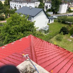 DACH-ROL ROLAND SZCZEPANIAK - Czerwony dach z dachówki ceramicznej po malowaniu, widok z góry, z aplikatorem natryskowym na pierwszym planie, w tle zabudowa jednorodzinna i zieleń.