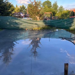 DACH-ROL ROLAND SZCZEPANIAK - Świeżo wylana, gładka posadzka z widocznym odbiciem drzew i nieba, zacierana mechanicznie, w tle zielona siatka maskująca i maszyny rolnicze.