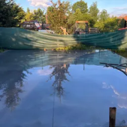 Świeżo wylana, gładka posadzka z widocznym odbiciem drzew i nieba, zacierana mechanicznie, w tle zielona siatka maskująca i maszyny rolnicze.