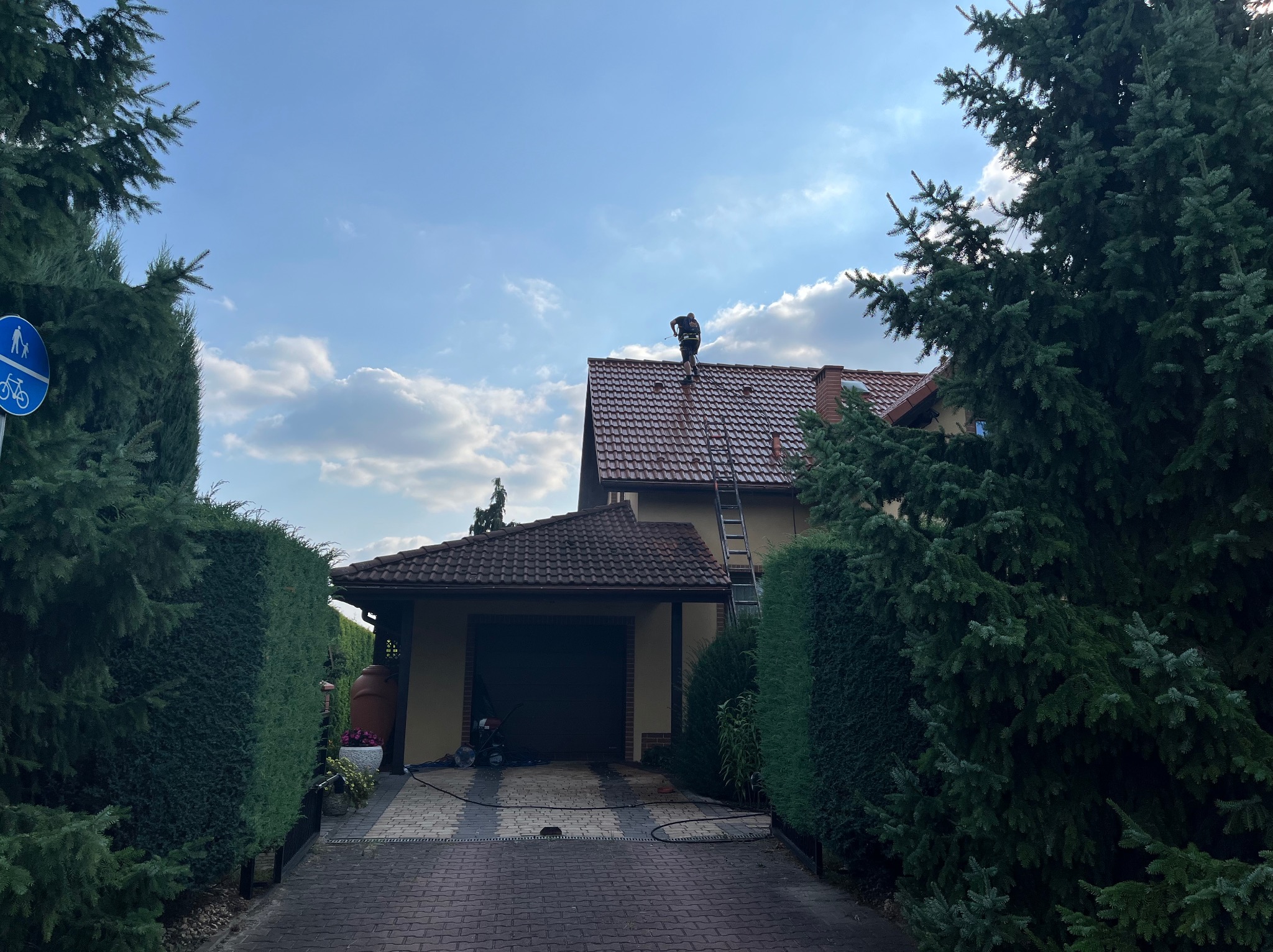 Pracownik na drabinie, na tle domu z brązową dachówką, wykonuje prace na dachu w słoneczny dzień.