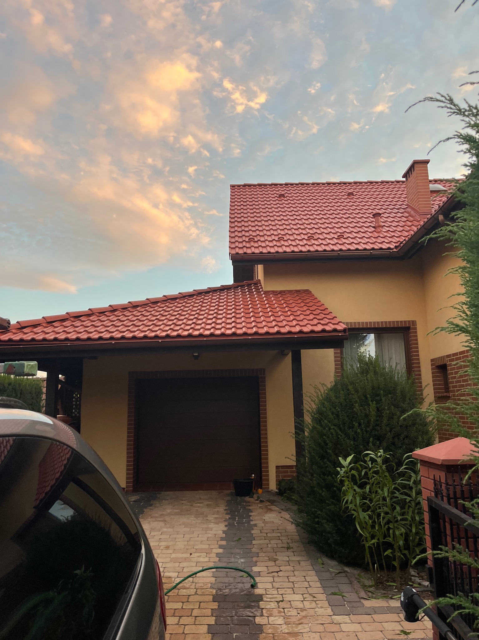 Dom jednorodzinny z nowym, czerwonym dachem z blachodachówki, widoczny fragment samochodu i ozdobny bruk podjazdu.
