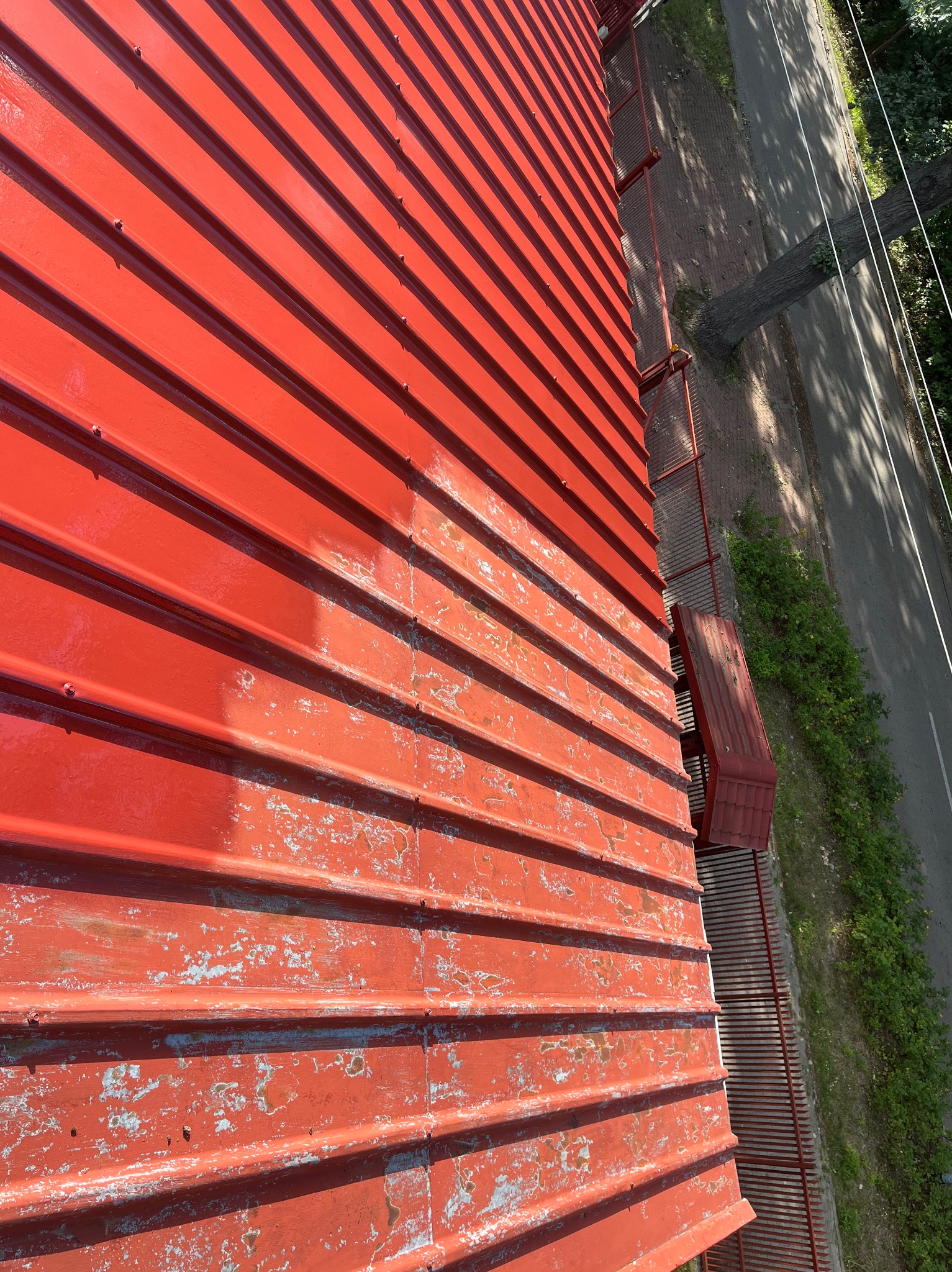 Czerwony, blaszany dach z widocznymi śladami odpryskującej farby, ujęcie z góry, widoczna krawędź dachu i fragment ulicy z zielenią.