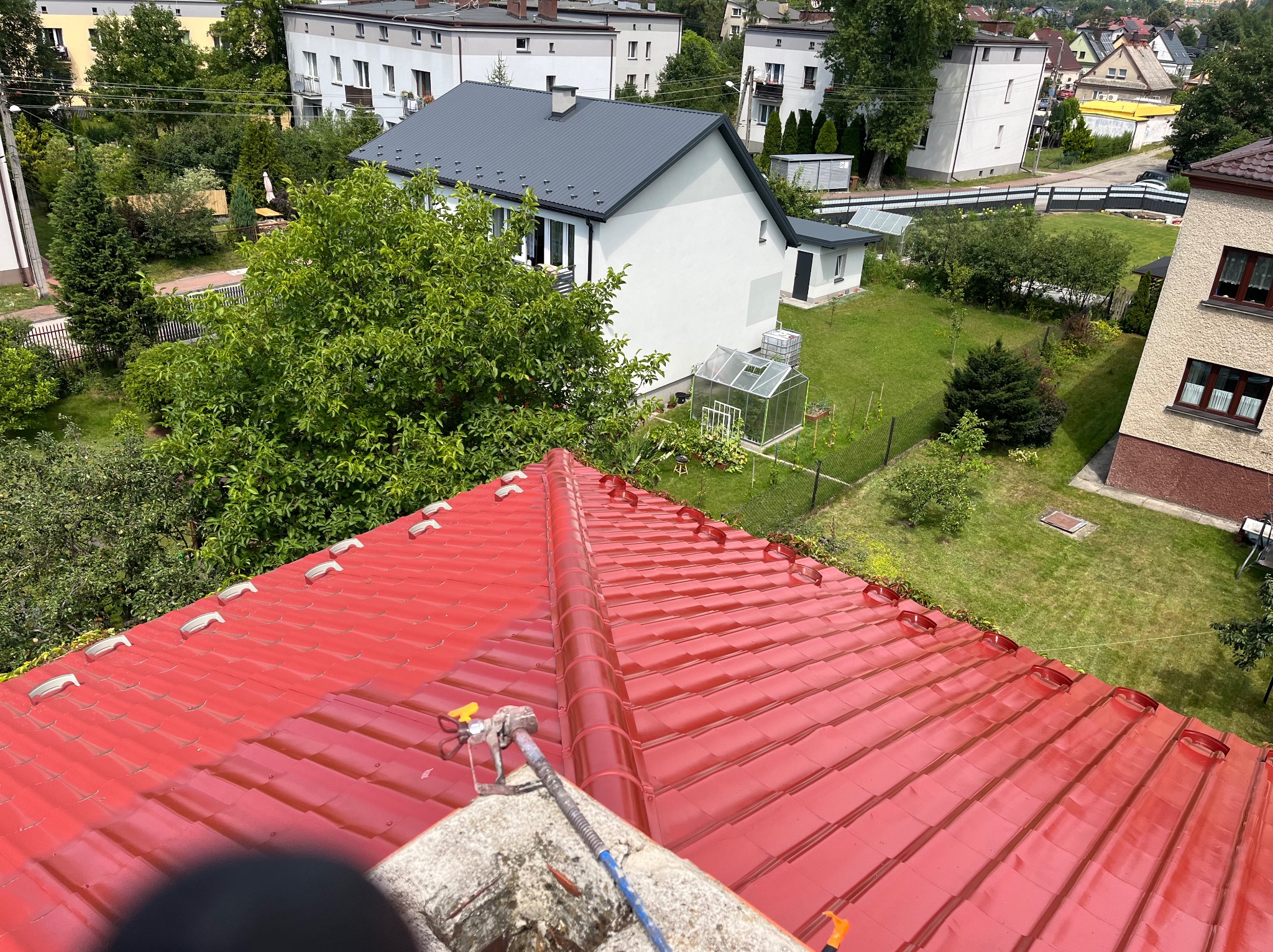 Czerwony dach z dachówki ceramicznej po malowaniu, widok z góry, z aplikatorem natryskowym na pierwszym planie, w tle zabudowa jednorodzinna i zieleń.