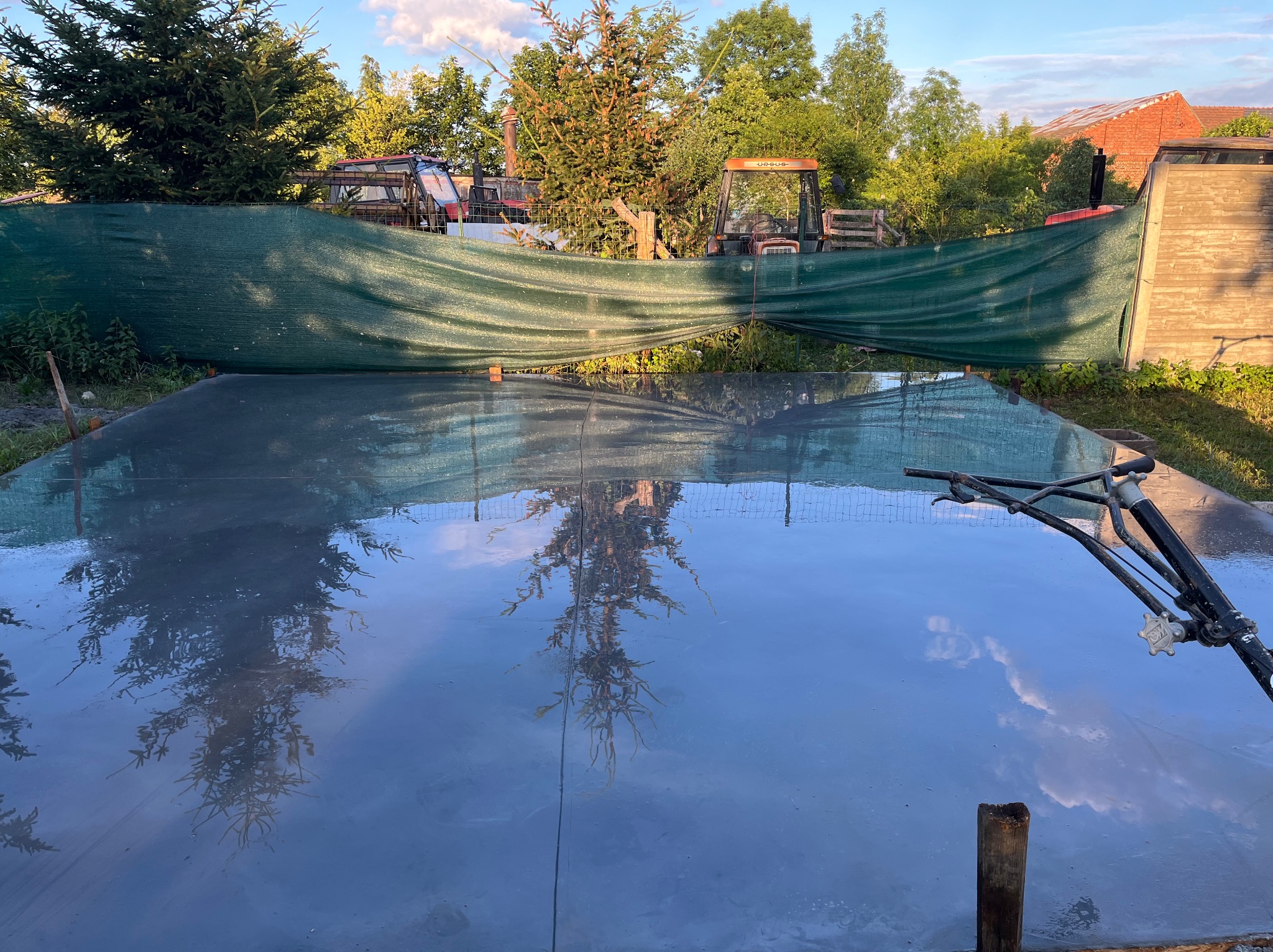 Świeżo wylana, gładka posadzka z widocznym odbiciem drzew i nieba, zacierana mechanicznie, w tle zielona siatka maskująca i maszyny rolnicze.