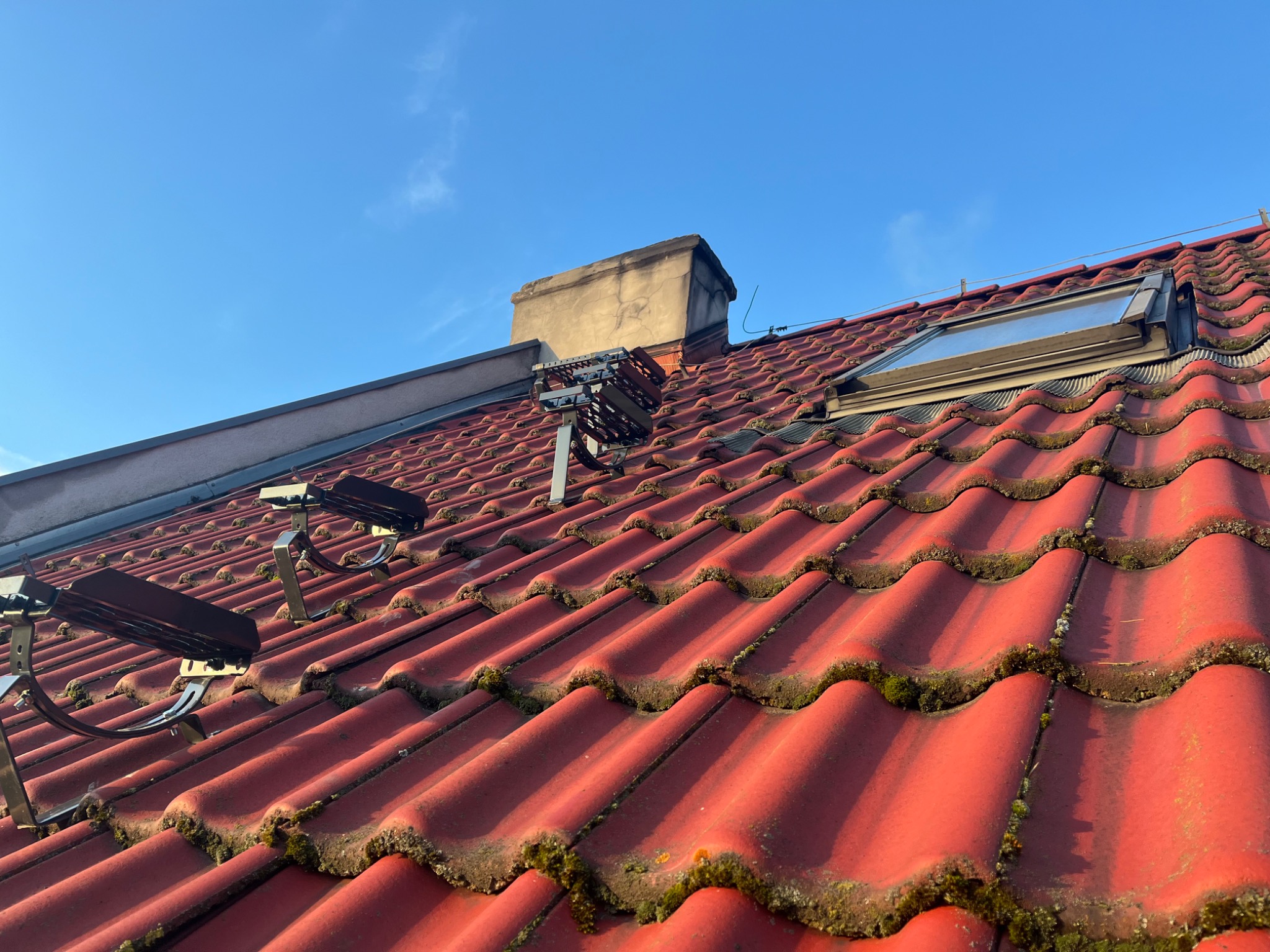 Czerwona dachówka ceramiczna z zamontowanymi stopniami kominiarskimi, oknem dachowym i kominem na tle błękitnego nieba.