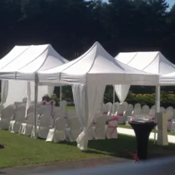 Białe namioty z draperiami i krzesłami przygotowane na uroczystość na trawniku w Katowicach, z widocznym stolikiem z kwiatami i kieliszkami.