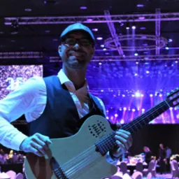 Uśmiechnięty muzyk w kamizelce i czapce trzyma gitarę Godin na tle sceny z niebieskimi światłami i ekranem z napisem '25'.