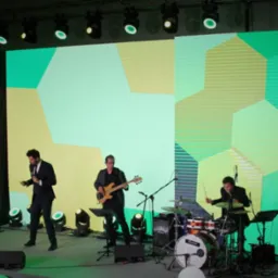 Zespół muzyczny w trakcie występu na scenie z geometrycznym tłem, oświetlony reflektorami.