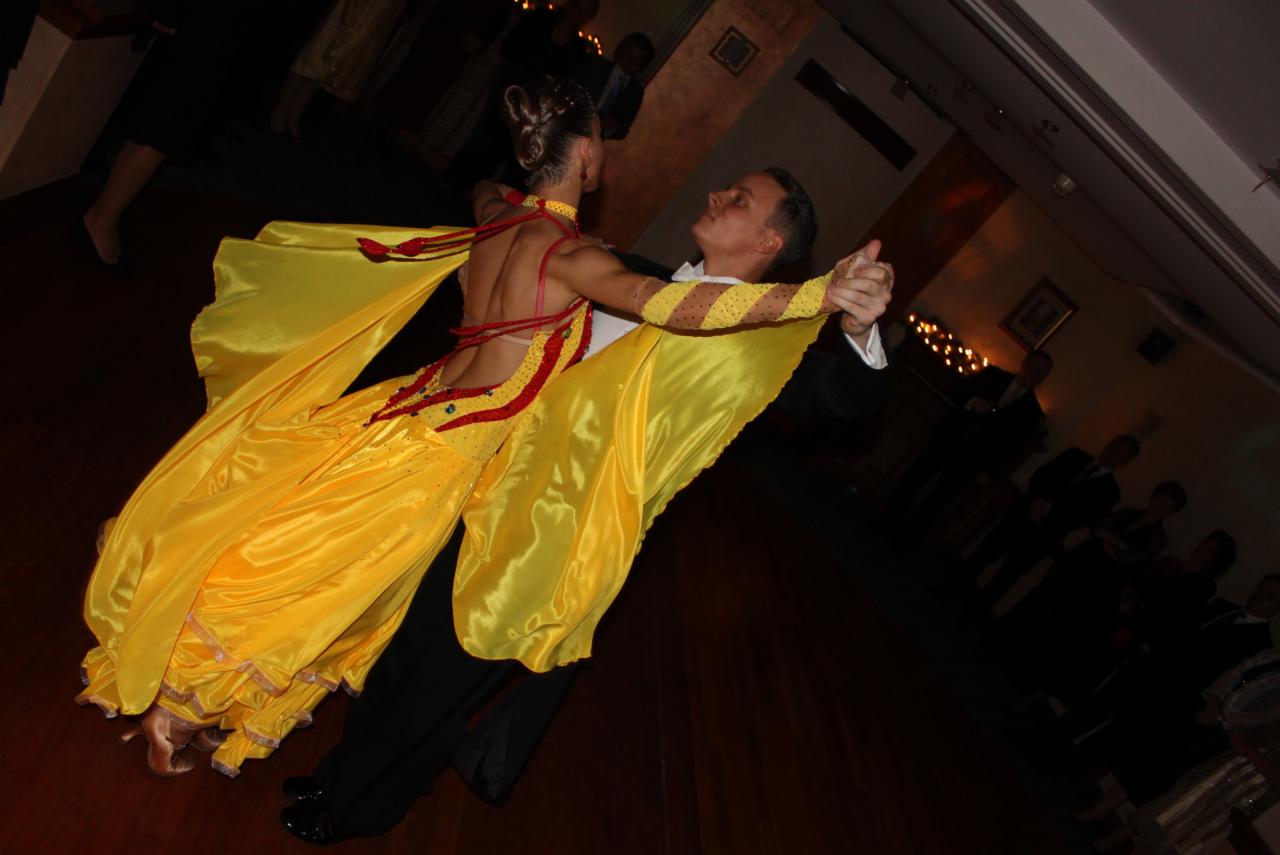 Para tancerzy w tańcu towarzyskim: mężczyzna w czarnym smokingu, kobieta w żółtej sukni z czerwonymi aplikacjami, parkiet i publiczność w tle.