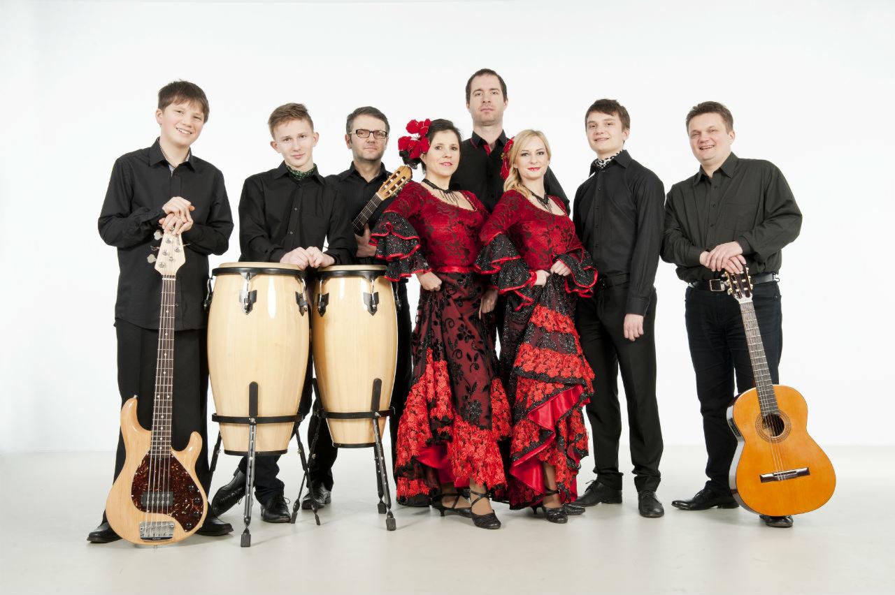 Zespół muzyczny w składzie siedmiu osób, w tym dwie wokalistki w czerwono-czarnych strojach flamenco, prezentujący instrumenty: gitara basowa, dwie congi i dwie gitary akustyczne, na białym tle.