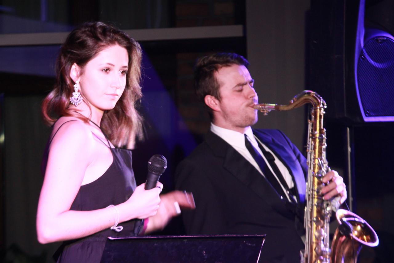 Wokalistka z mikrofonem i saksofonista w garniturze występują na scenie podczas imprezy, oświetleni fioletowym światłem.