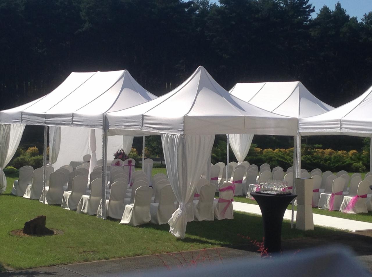 Białe namioty z draperiami i krzesłami przygotowane na uroczystość na trawniku w Katowicach, z widocznym stolikiem z kwiatami i kieliszkami.