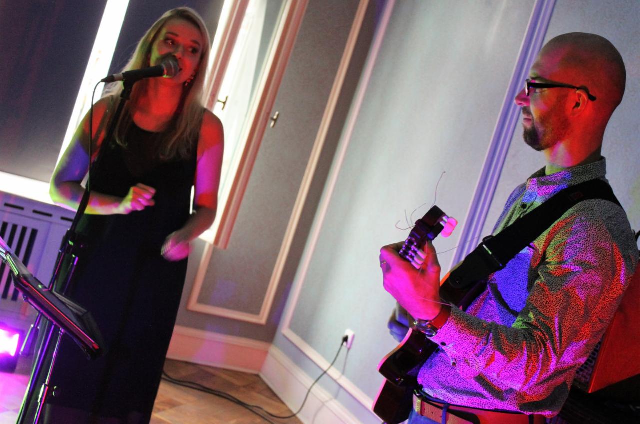 Wokalistka z mikrofonem i gitarzysta z ukulele występują na tle klasycznego wnętrza z boazerią, oświetleni kolorowymi światłami.