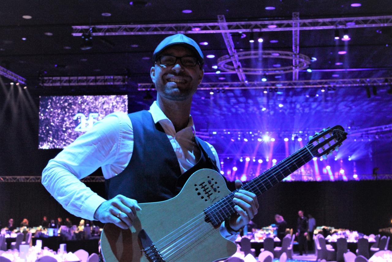 Uśmiechnięty muzyk w kamizelce i czapce trzyma gitarę Godin na tle sceny z niebieskimi światłami i ekranem z napisem '25'.