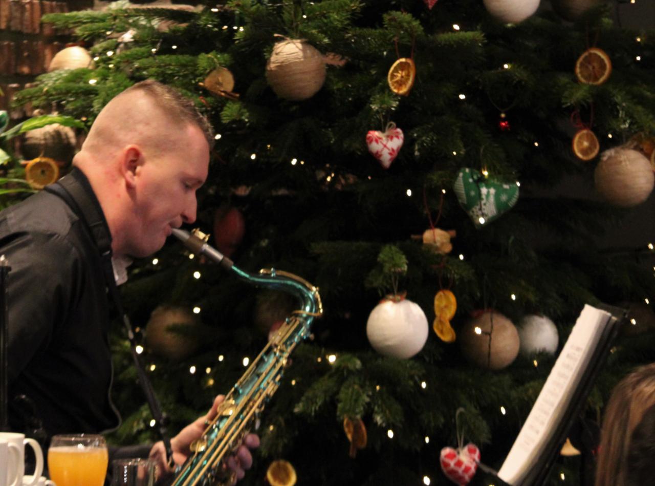 Saksofonista gra kolędy przed choinką z ozdobami DIY z suszonych pomarańczy i serduszek z materiału.