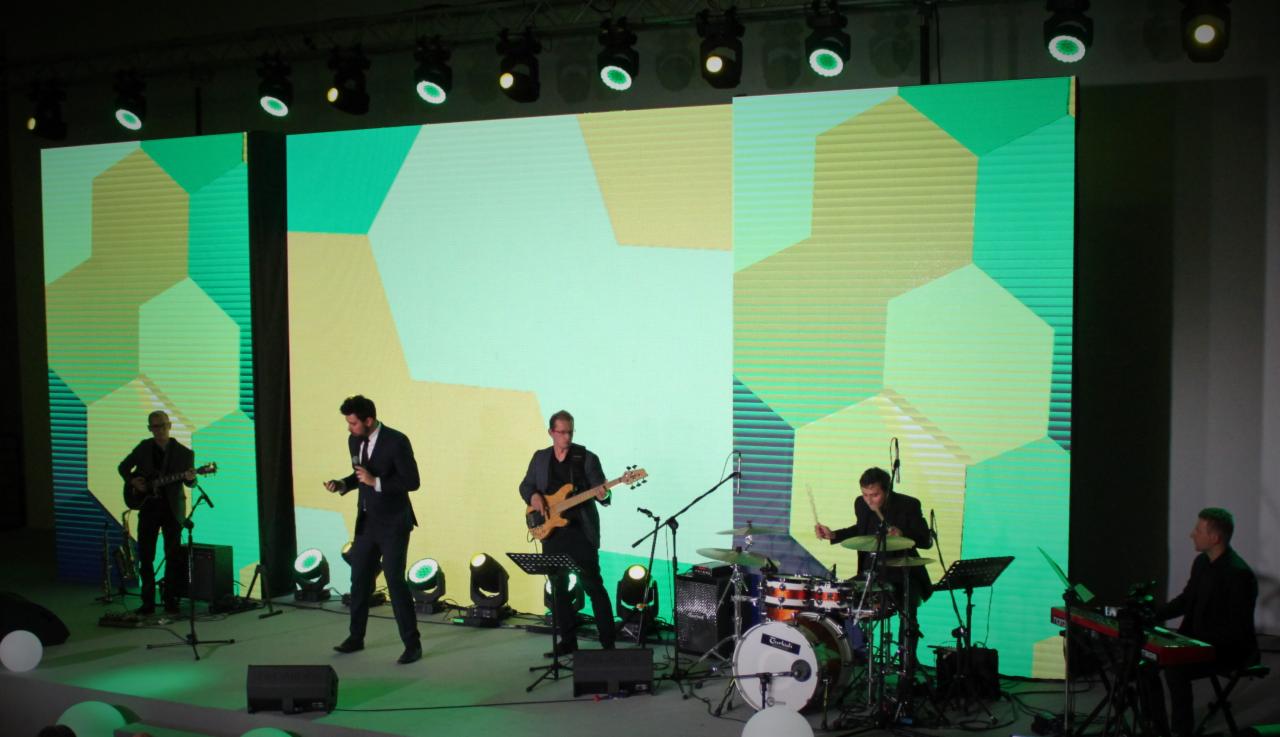 Zespół muzyczny w trakcie występu na scenie z geometrycznym tłem, oświetlony reflektorami.