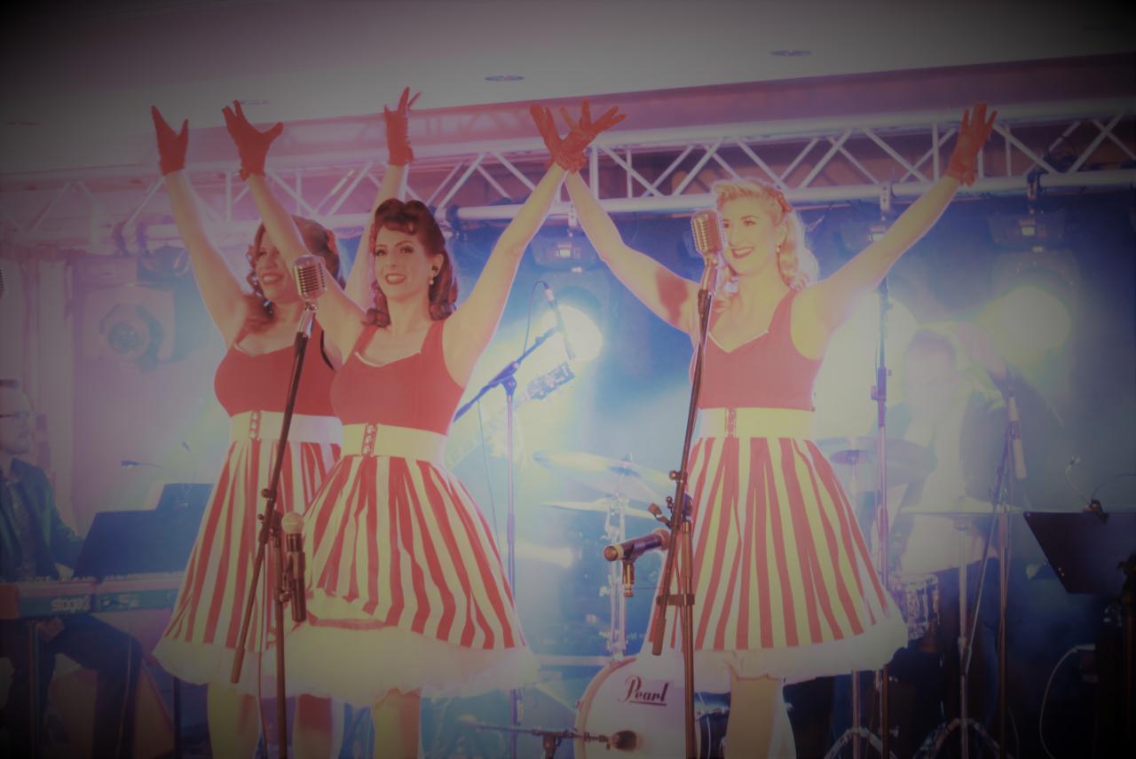 Trzy wokalistki w czerwono-białych sukienkach retro z podniesionymi rękami występują na scenie z mikrofonami w stylu vintage, widoczny perkusista w tle.